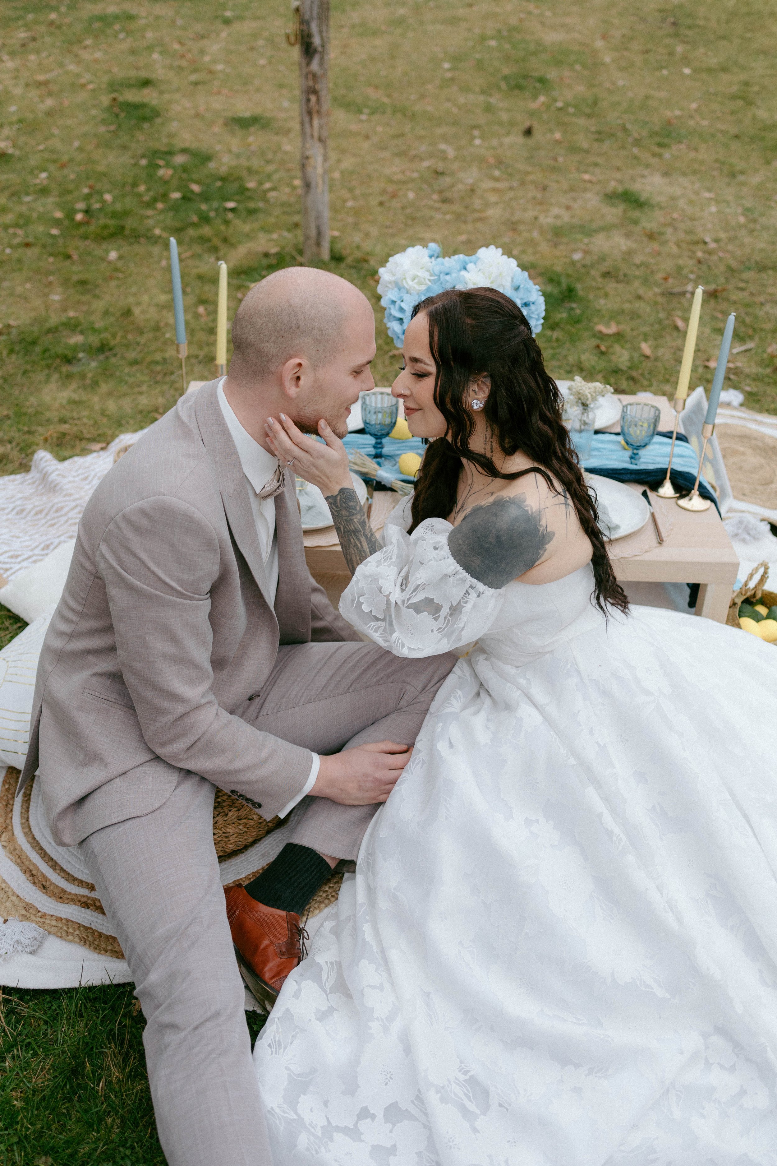 A couple on their wedding day, sitting on a blanket in a grassy outdoor setting, smiling and looking at each other, with decorated table and candles in the background.