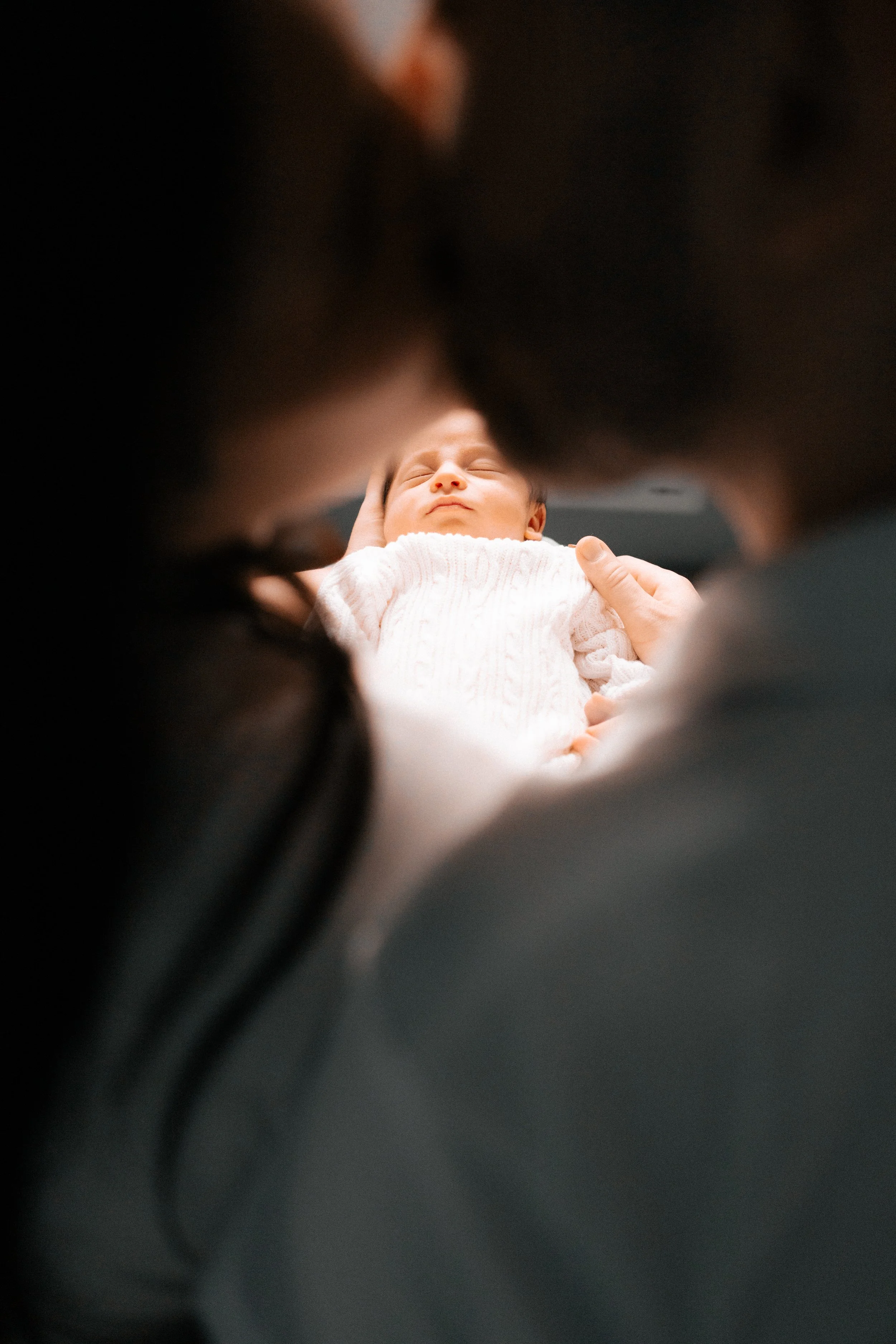 A person holding a baby lying on their back, wrapped in a white blanket, with a neutral expression and eyes closed.