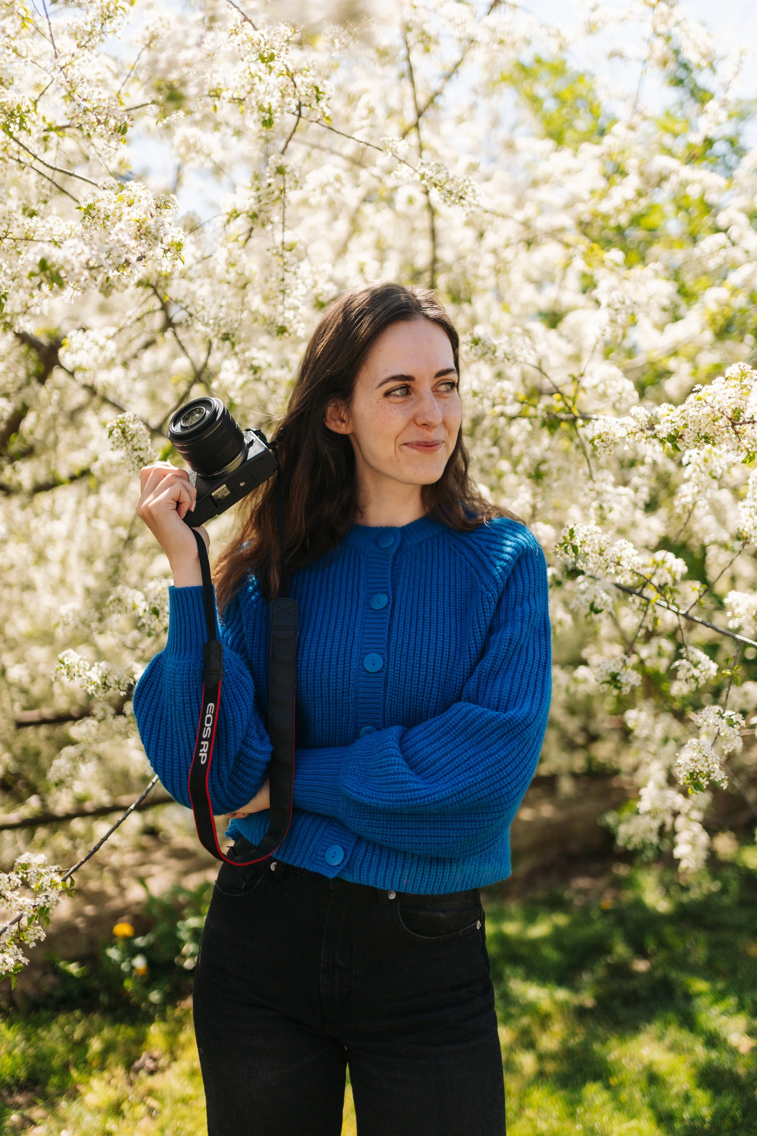 A woman with long brown hair wearing a blue sweater and black pants stands outdoors among blooming white flowers and trees, holding a camera over her shoulder.