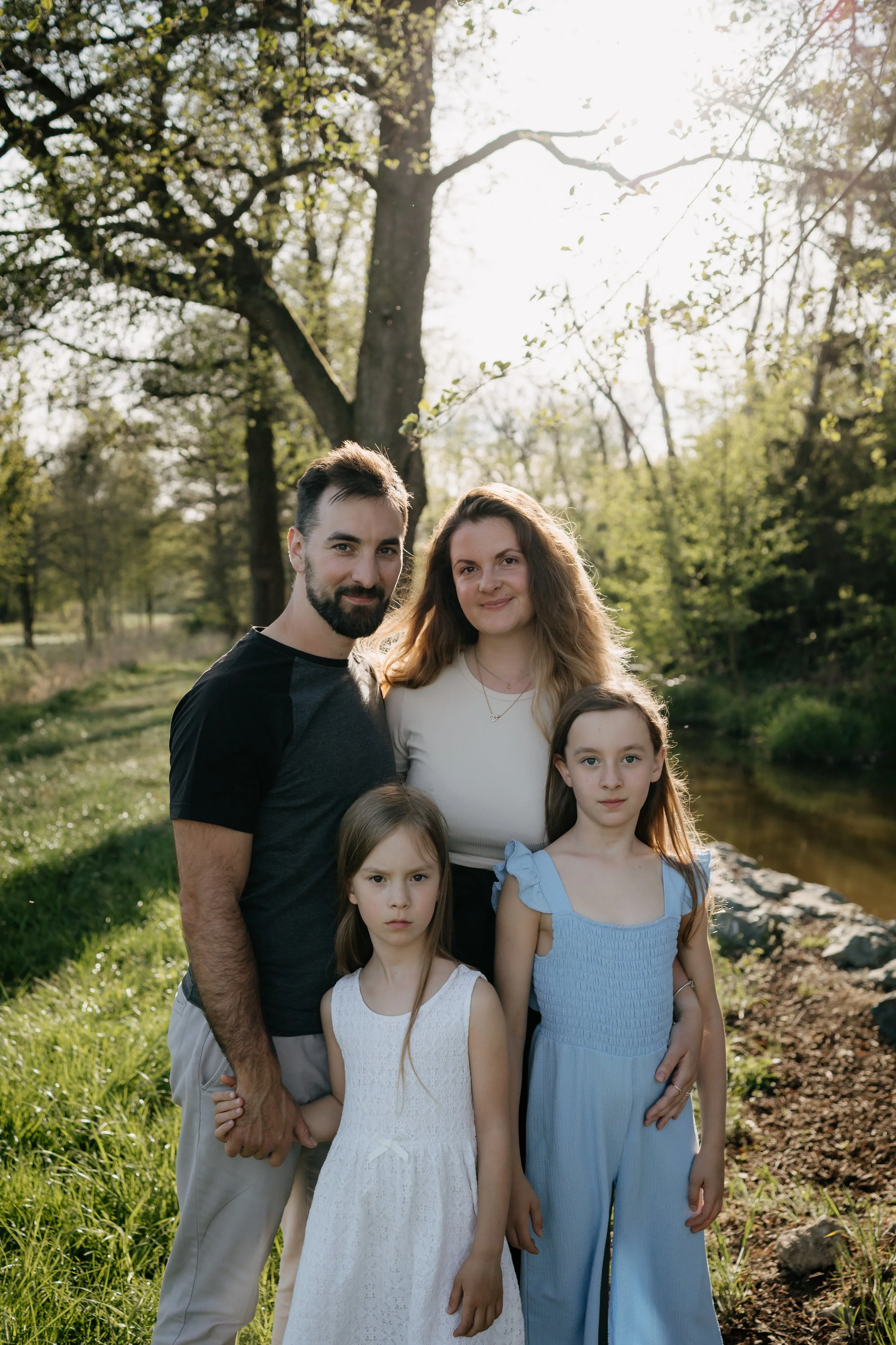 A family of five standing outdoors near a river, with trees and sunlight in the background. The father has dark hair and a beard, wearing a black and gray shirt. The mother has wavy, light brown hair, wearing a white top. Two young girls are in front