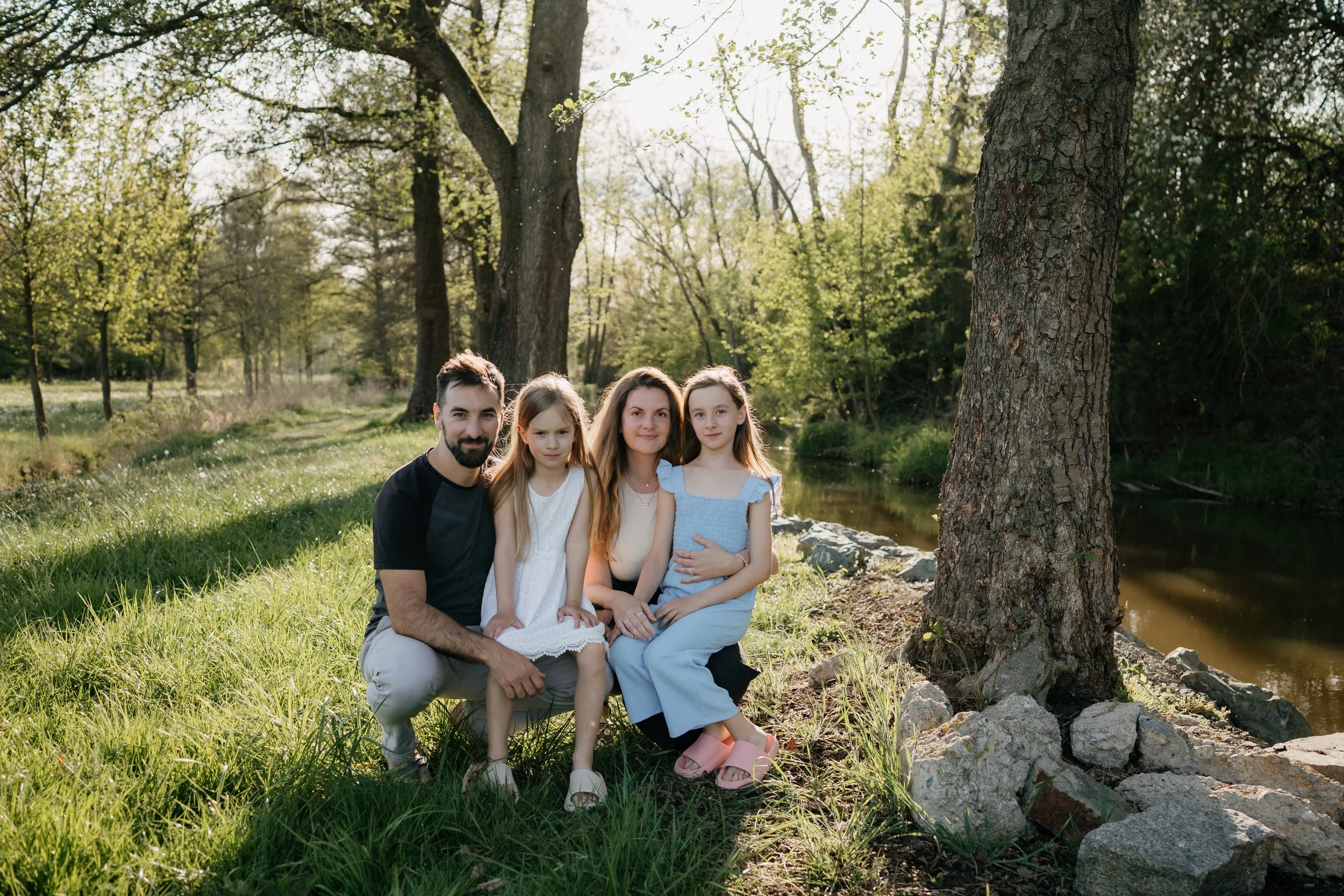 A family of five sitting outdoors on grass near a tree and stream in a wooded area during daylight.