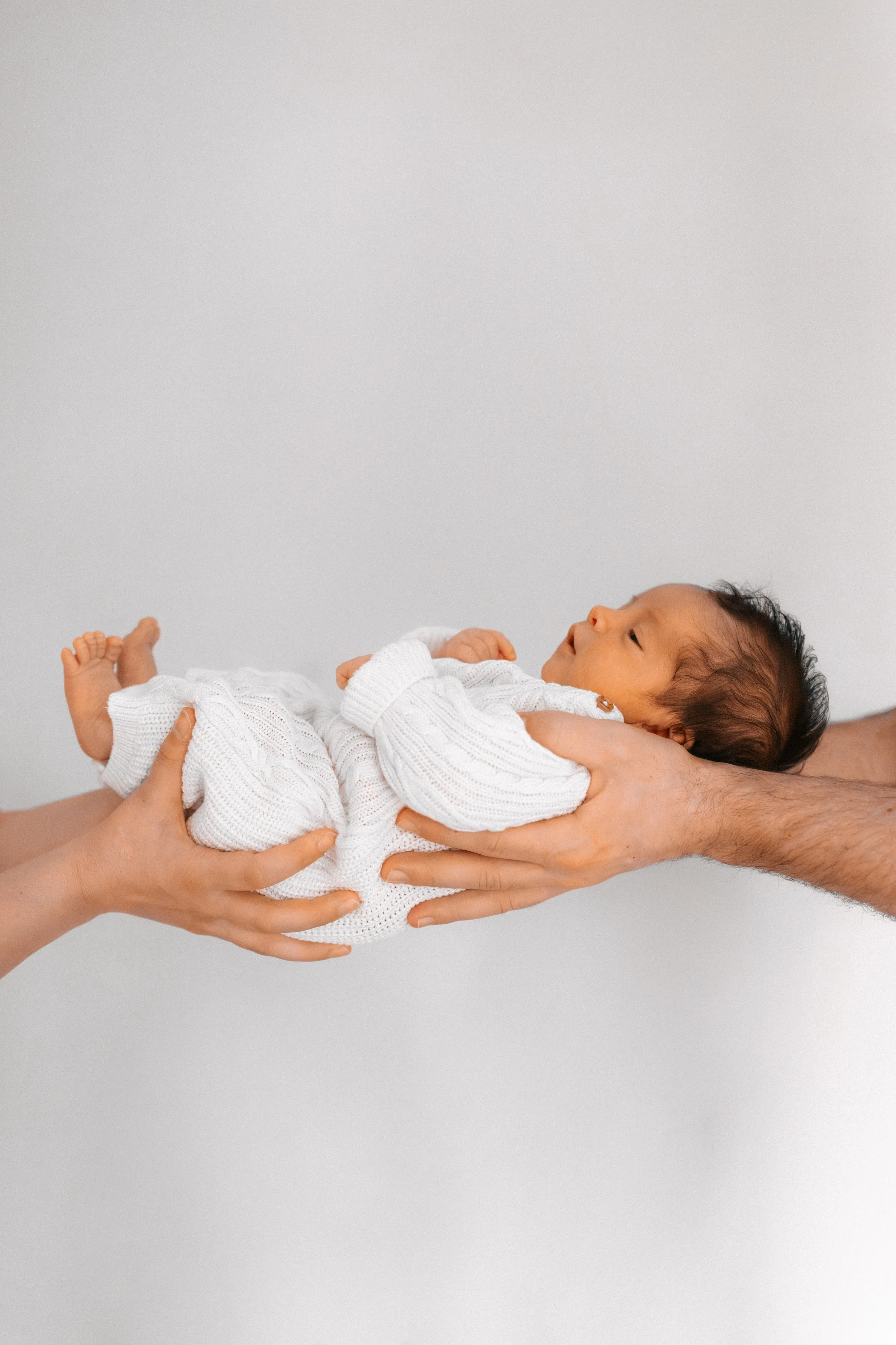 A newborn baby wrapped in a white knit blanket being gently held by multiple hands against a plain background.