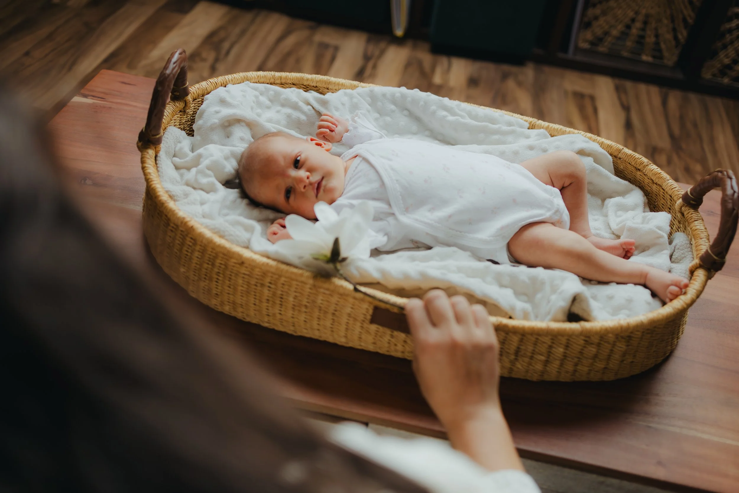 Baby lying on a wicker bassinet with white blankets, looking at the camera with a calm expression.