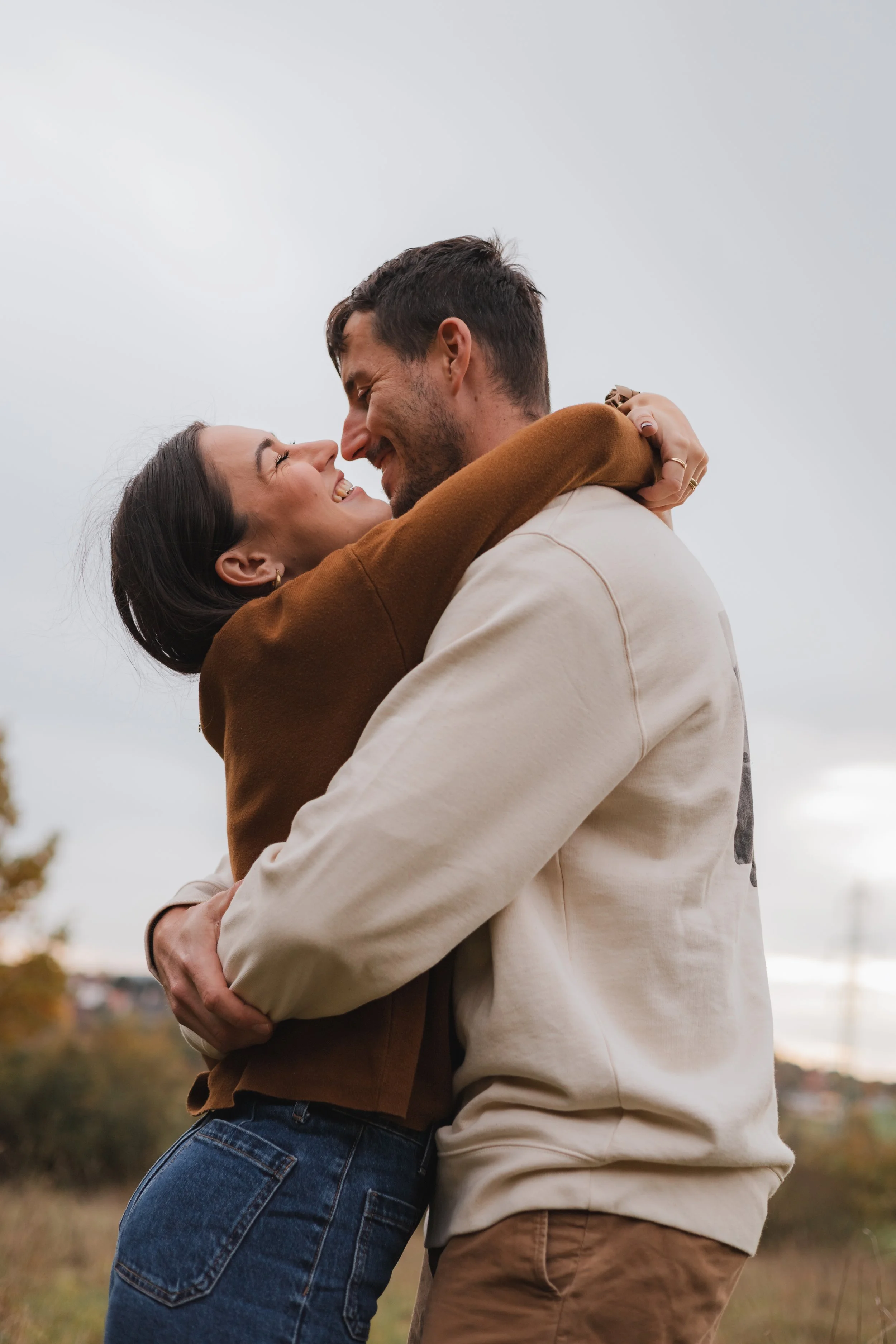A couple hugging outdoors on a cloudy day, smiling and looking into each other's eyes.