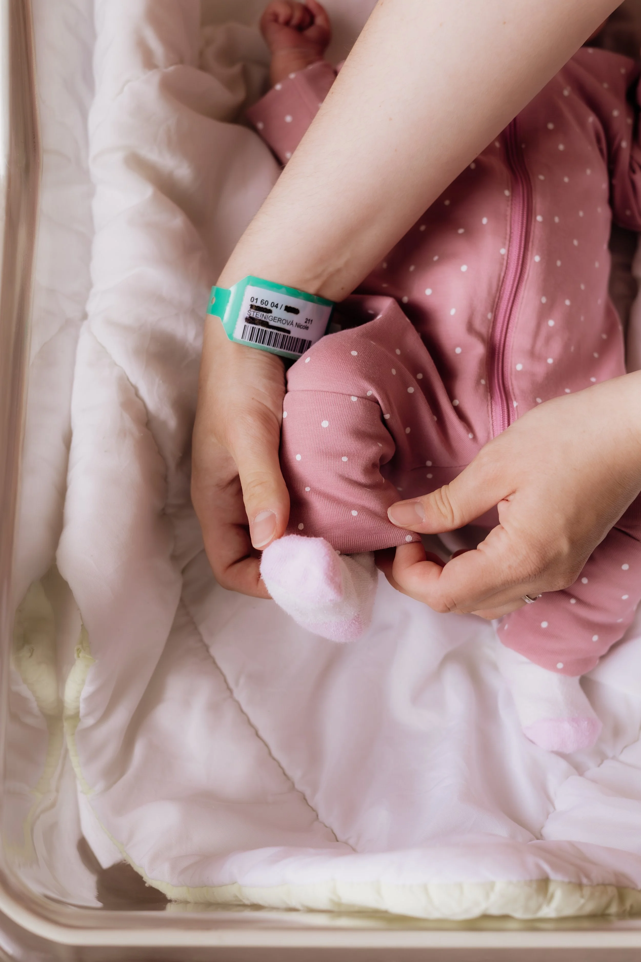 Newborn baby lying in a hospital bassinet, wearing pink polka dot clothing, with an identification bracelet on the wrist of an adult supporting them.