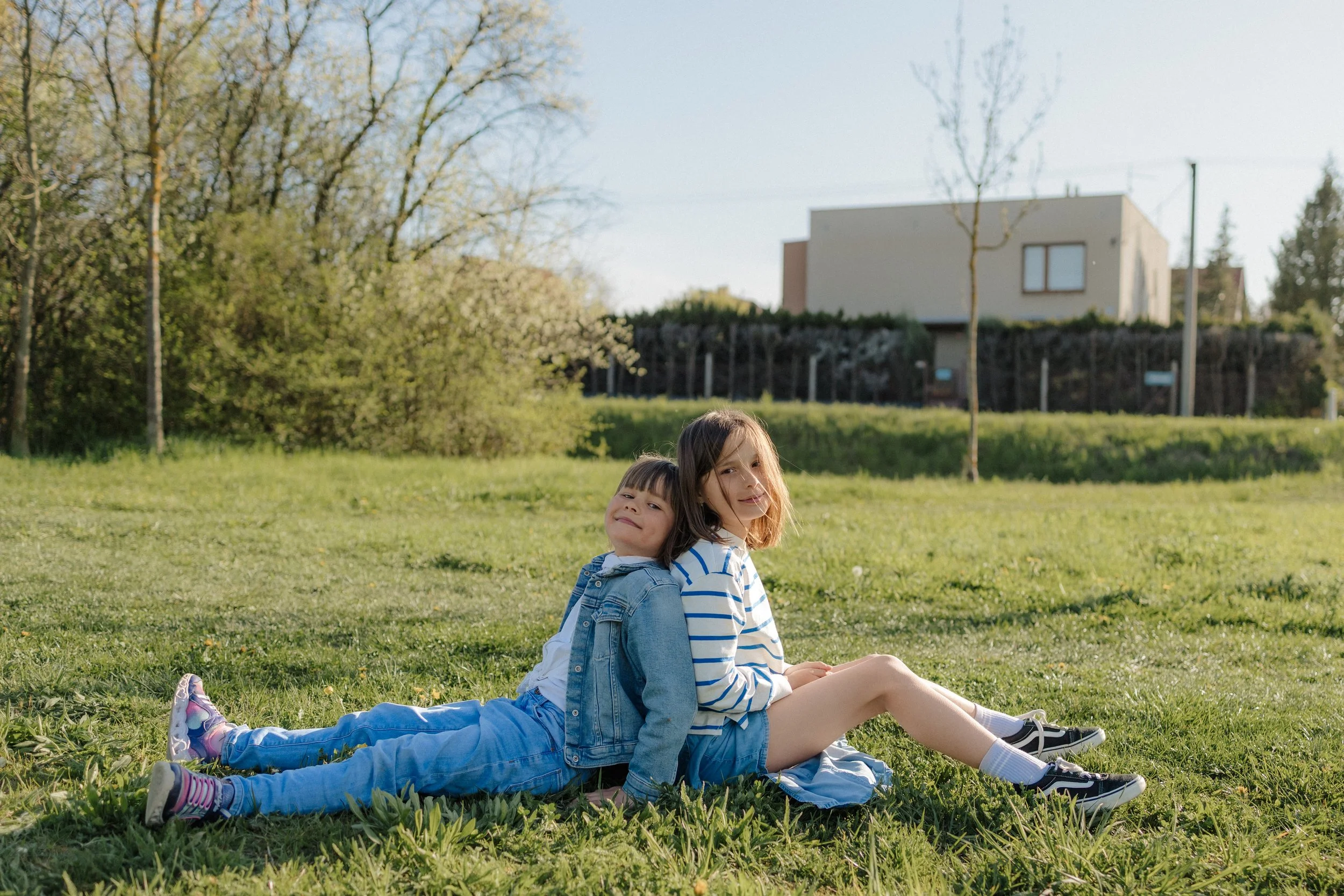 Two kids sitting on a grassy field, leaning against each other with a backdrop of leafless and budding trees and a modern house under a clear, bright sky.