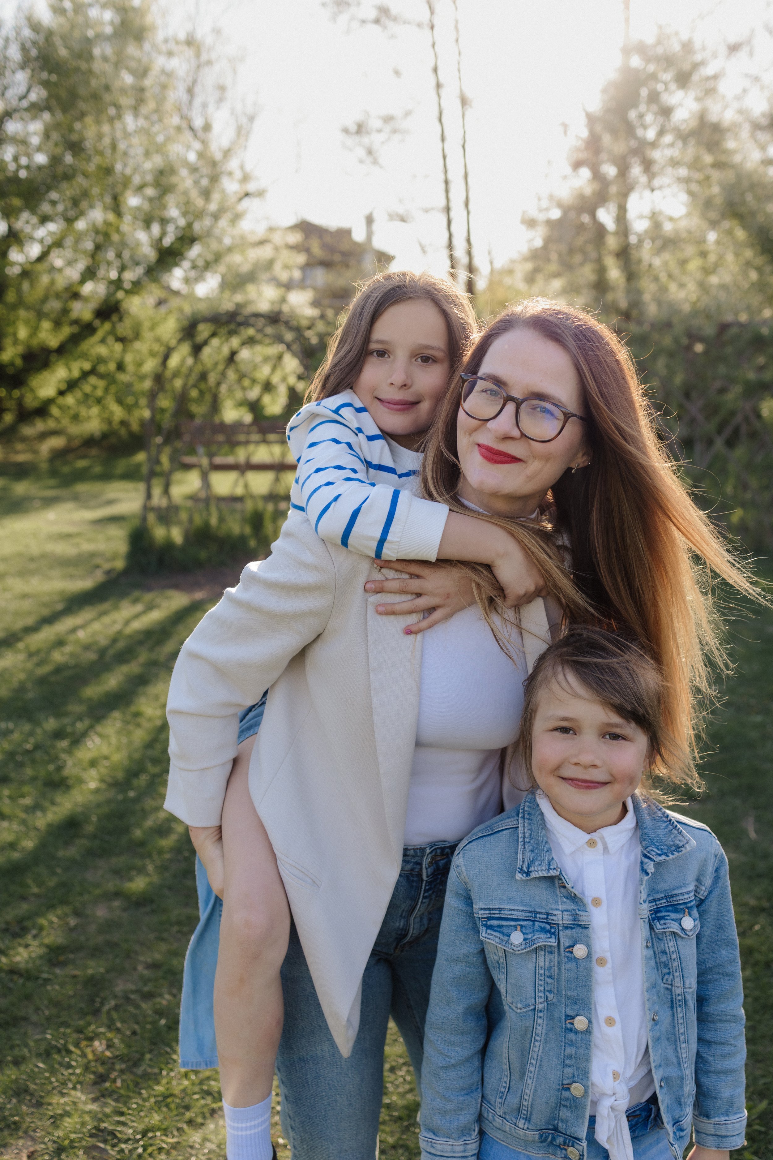 A woman with glasses and long reddish hair smiles as she holds a girl on her back, and there is a boy standing in front of them outdoors in a park with greenery and sunlight.