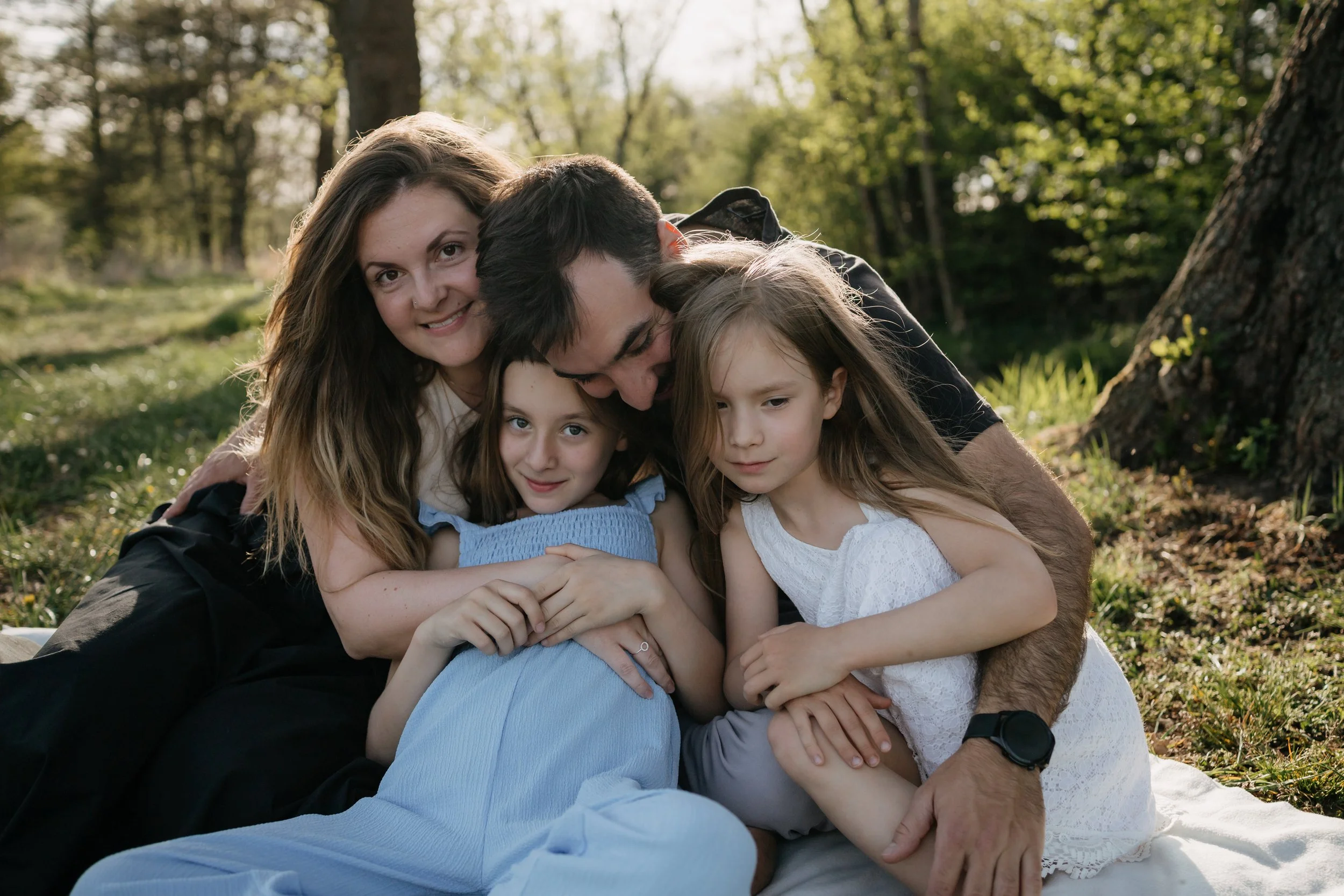 A woman, two young girls, and a young man hug and smile outdoors in a green, wooded area during daytime.