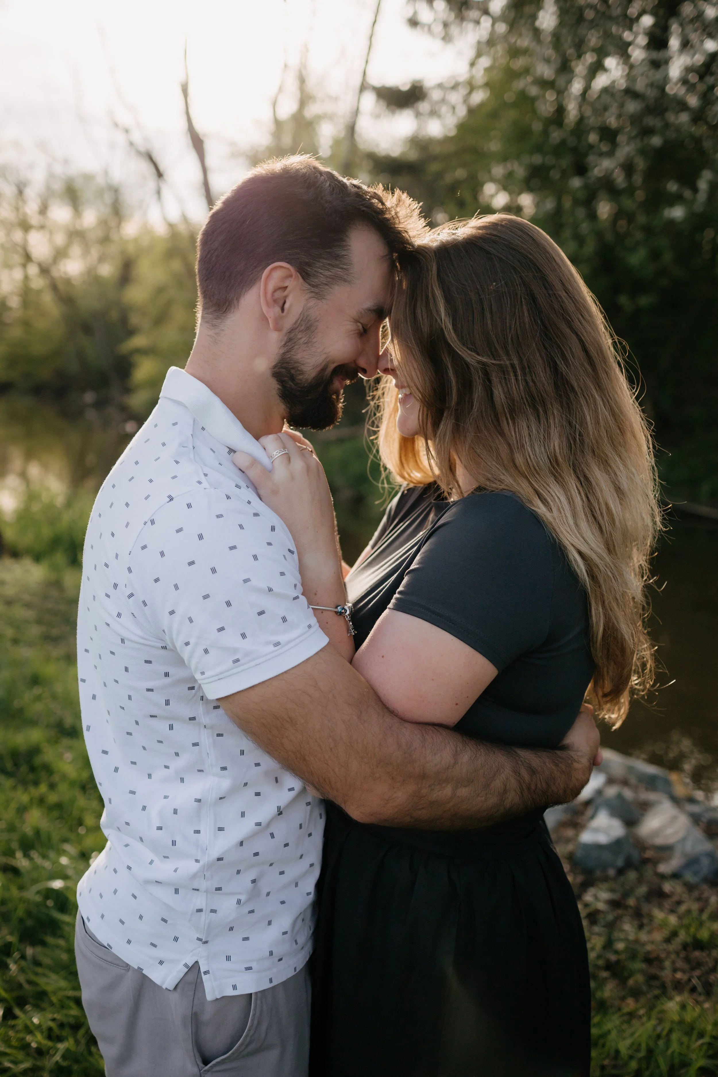 A couple embracing outdoors near a river with trees in the background, their foreheads touching and eyes closed, smiling.