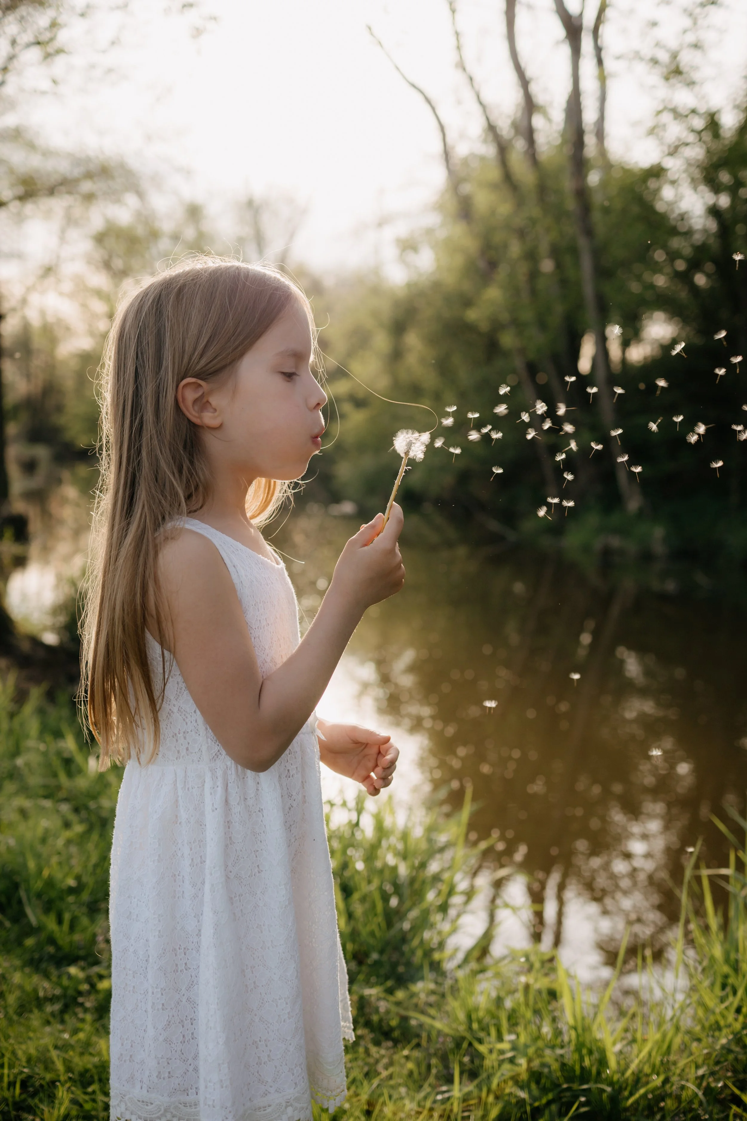 A young girl with long light brown hair in a white dress blowing on a dandelion near a body of water with trees in the background.