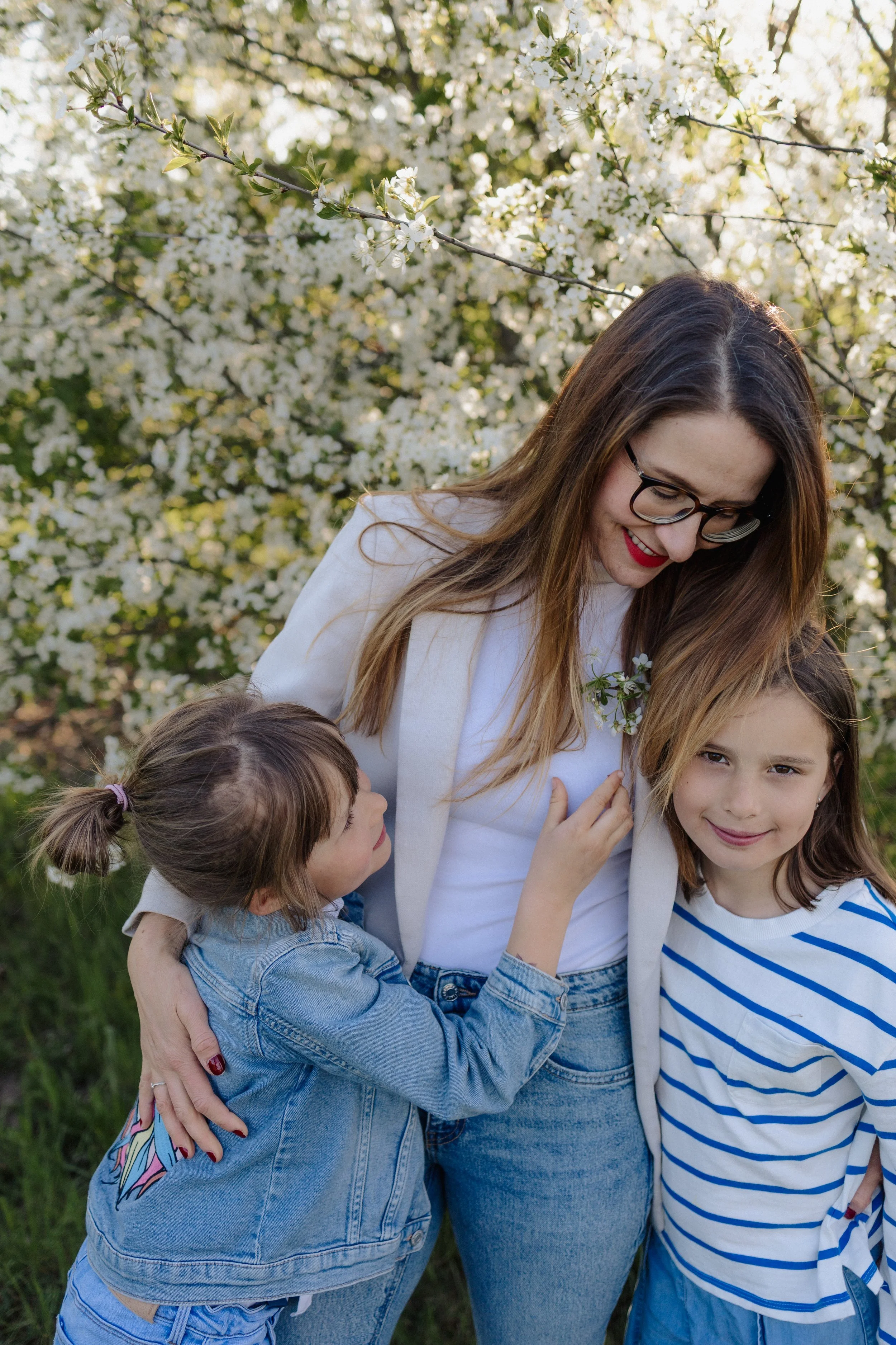 A woman with glasses and red lipstick smiling surrounded by children outdoors with blossoming trees in the background.