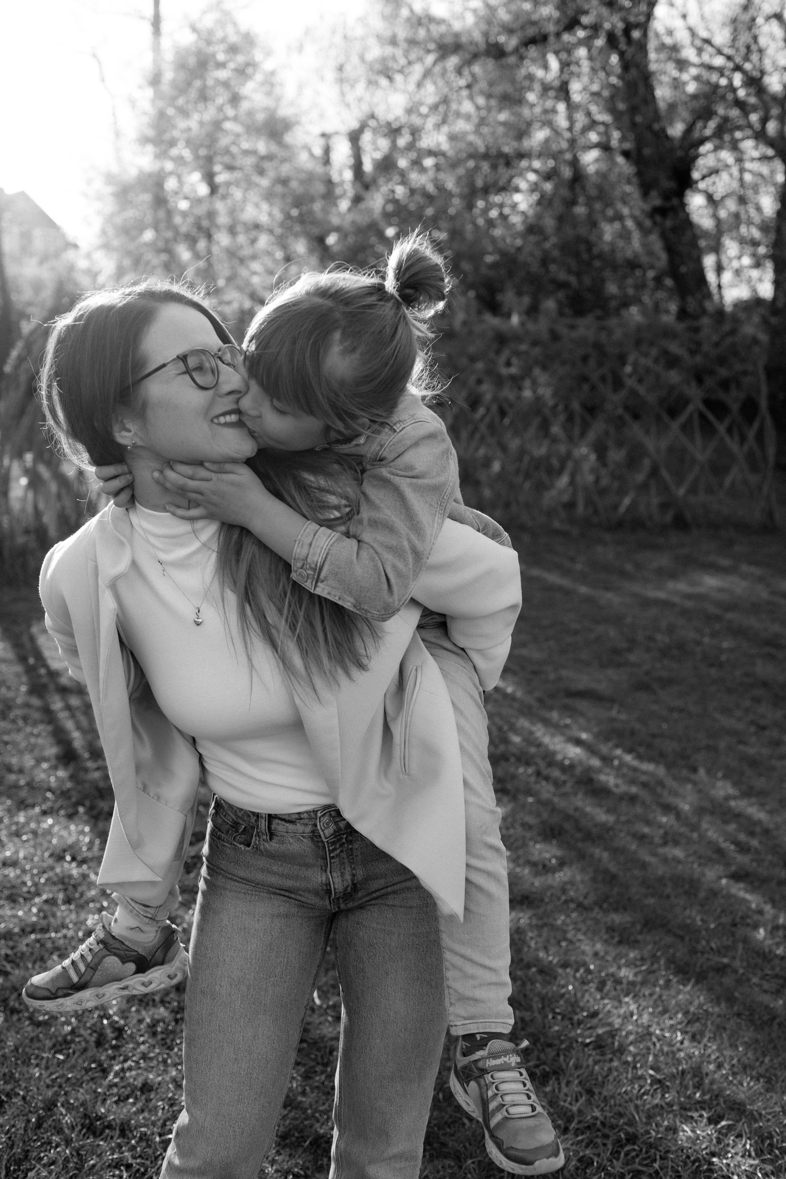 A woman holding a girl in the outdoors during daytime, both smiling and sharing a hug, with trees and greenery in the background.