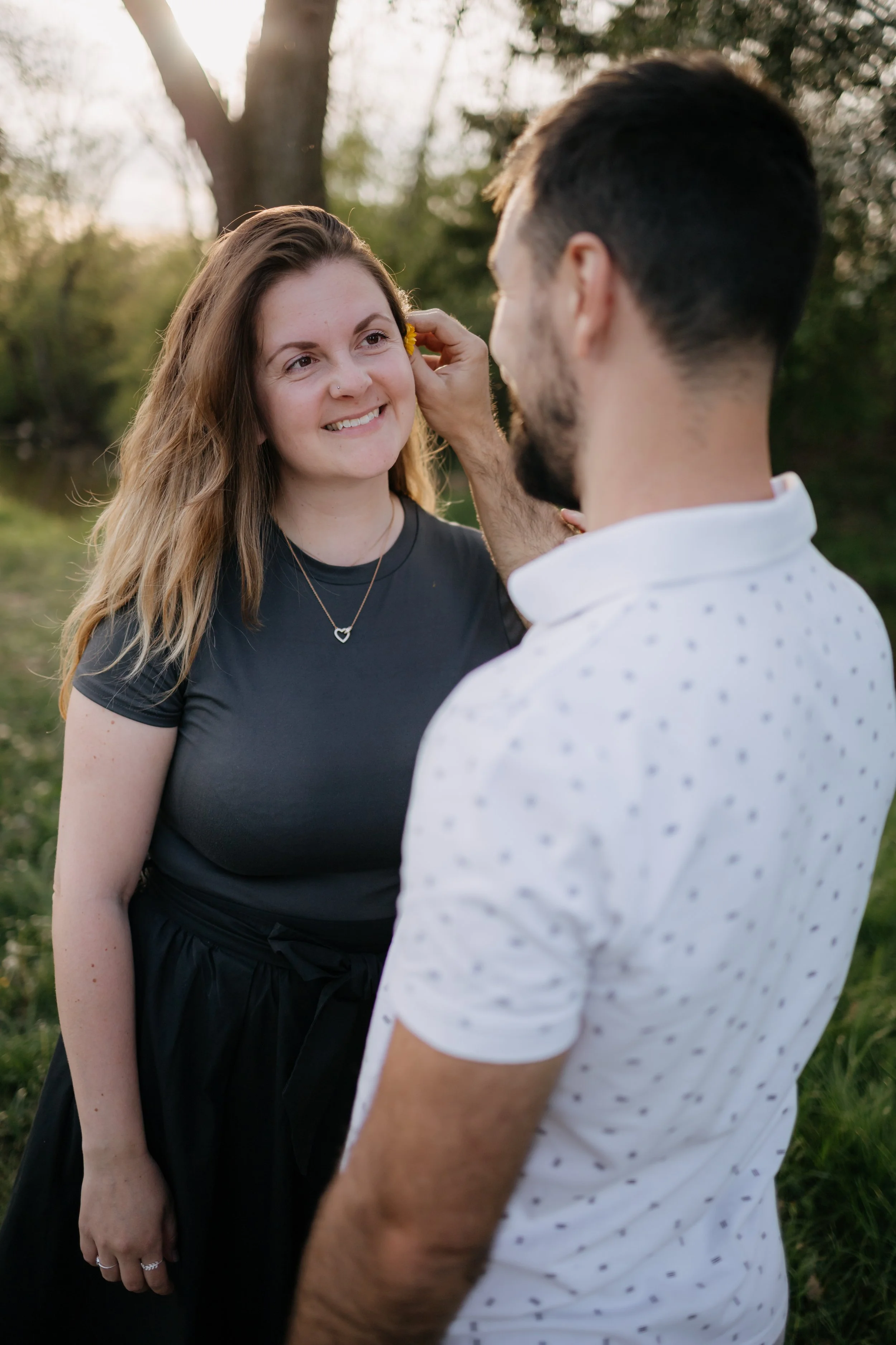 A man and woman outdoors with trees in the background. The woman is smiling, with long wavy hair, wearing a black top and a necklace with a heart pendant. The man, whose face is not visible, is touching the woman's face, wearing a white shirt with sm