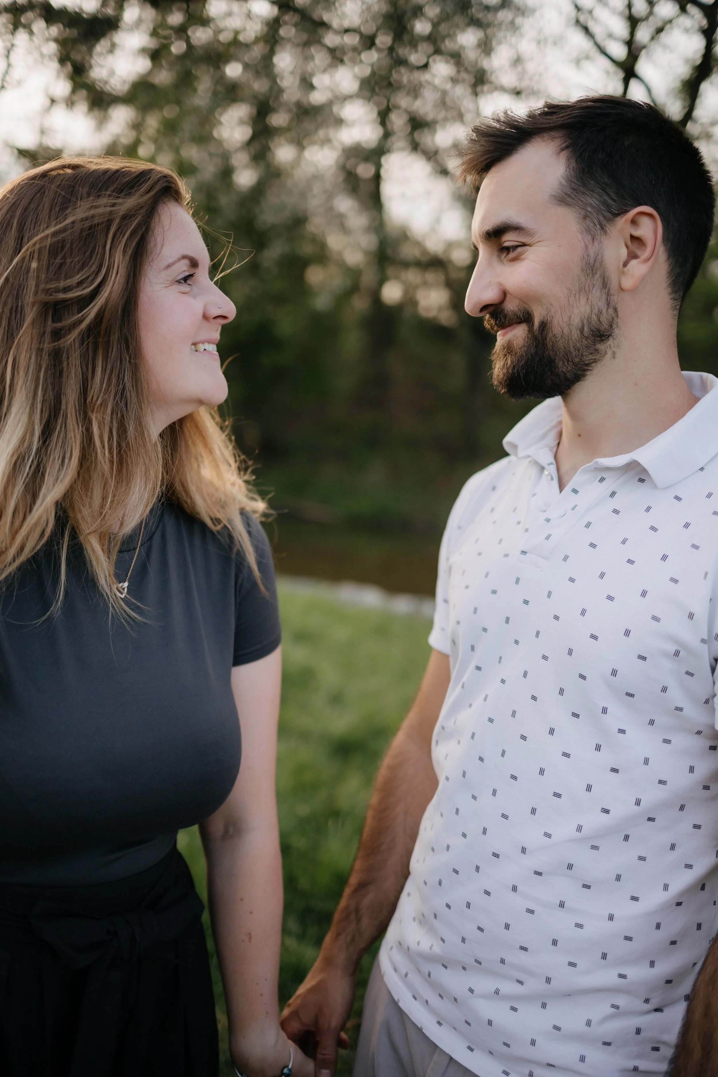 A woman and a man holding hands outdoors, looking at each other and smiling, with trees in the background.
