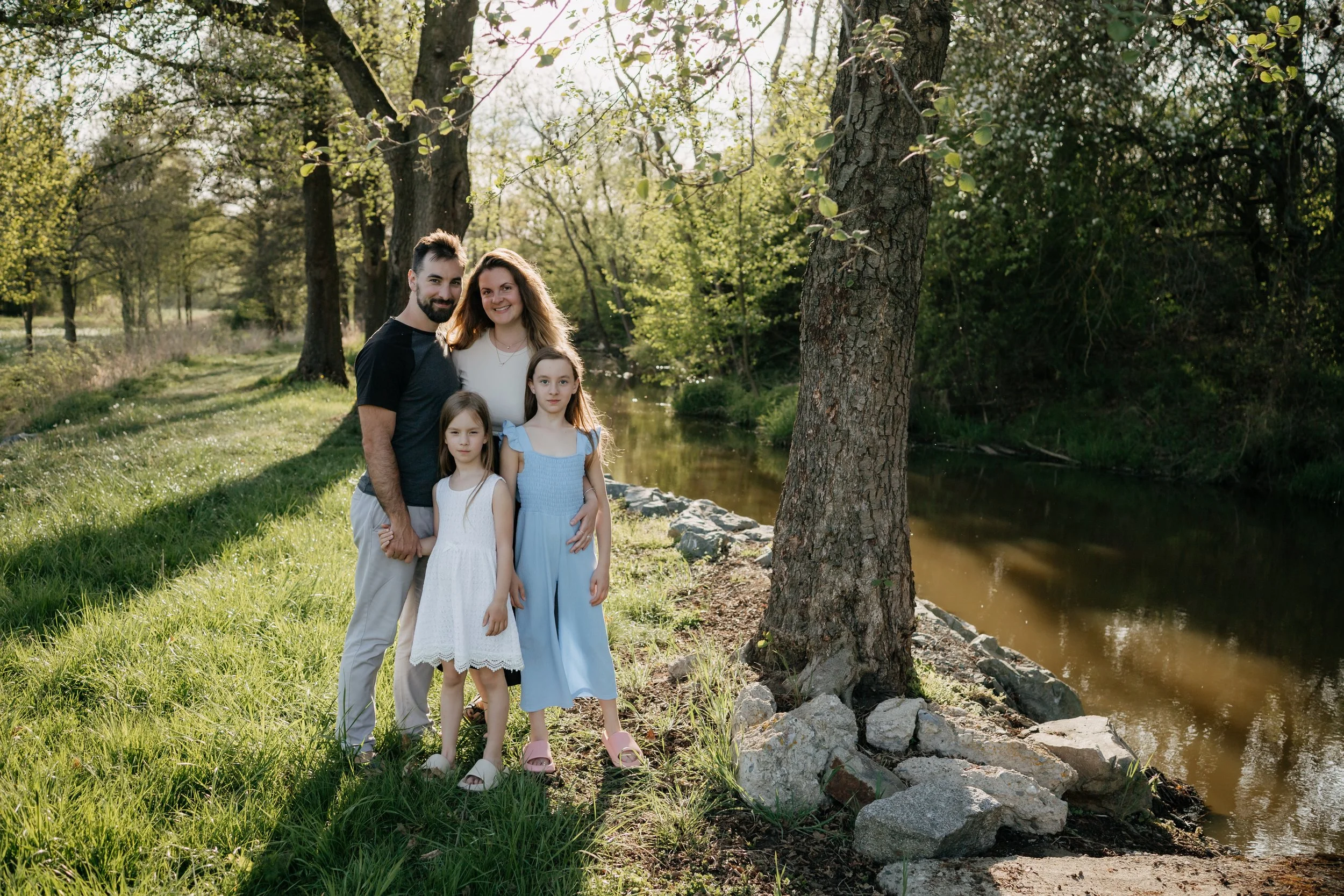 Family of four standing by a river in a park with trees in the background, smiling at the camera.