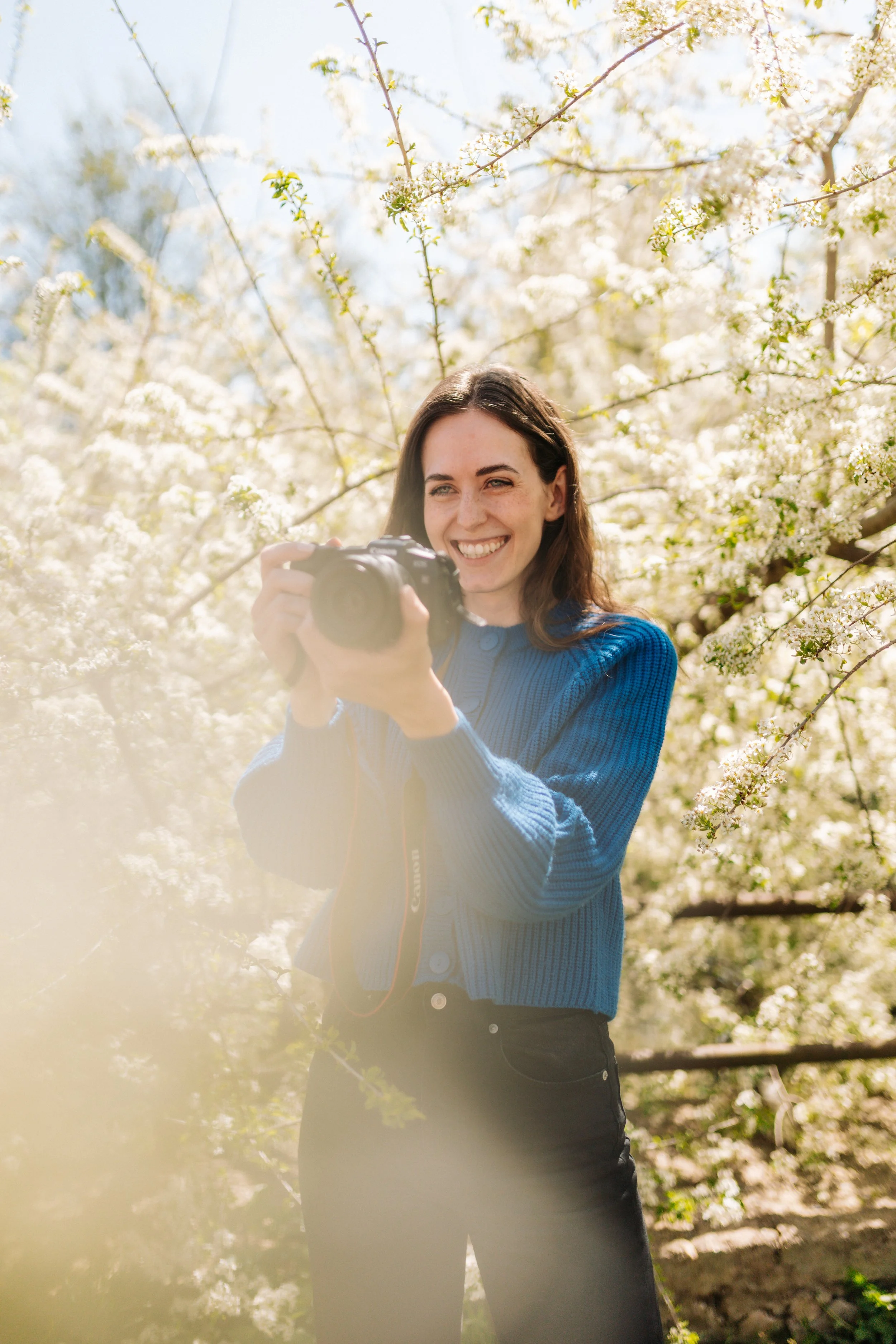 A woman with long brown hair smiling and holding a camera, standing outdoors among blooming white flowers on trees.