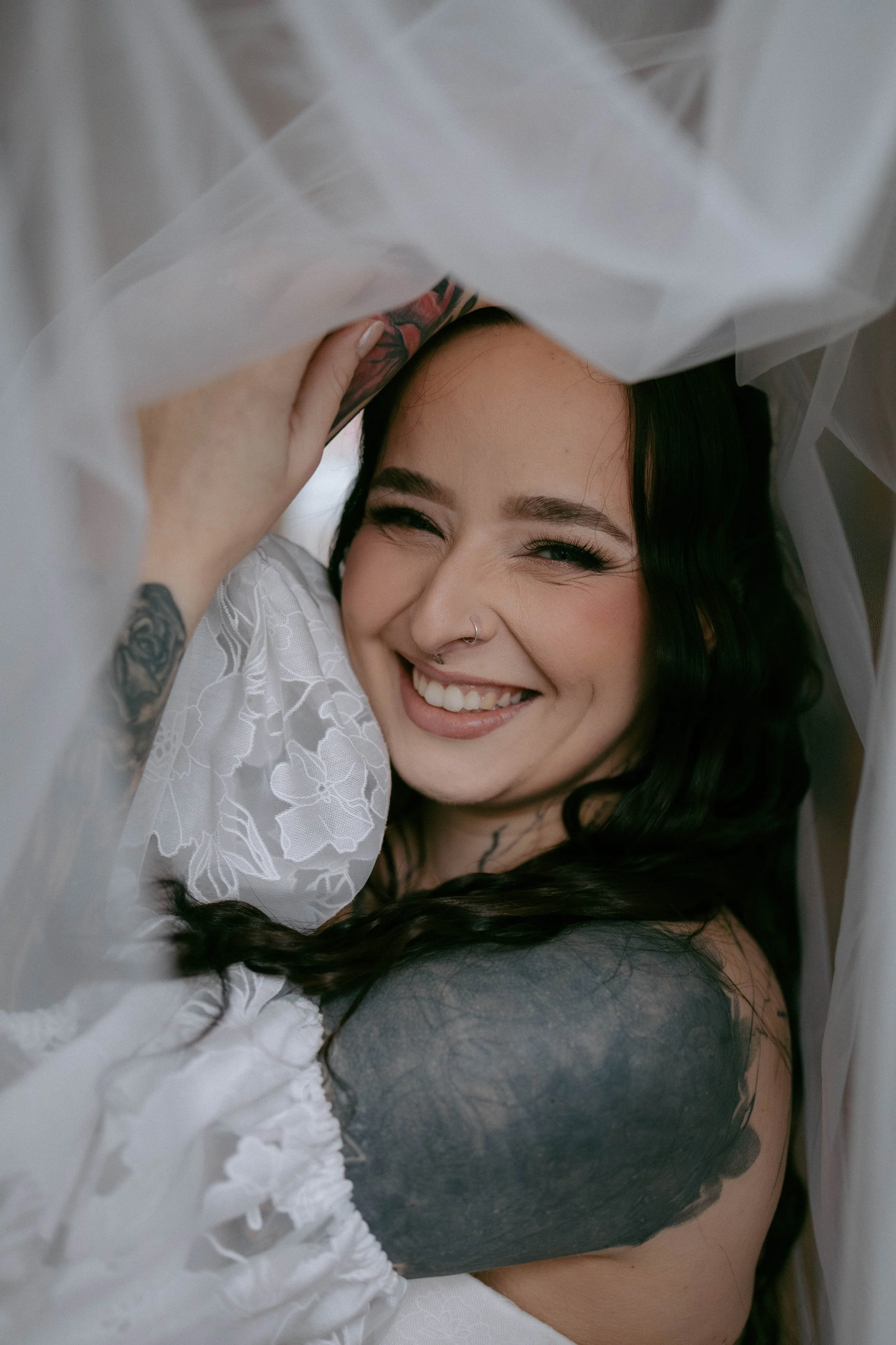 A woman with dark hair, smiling and squinting, lying beneath white, semi-transparent fabric with floral embroidery, revealing a tattoo on her arm and shoulder.