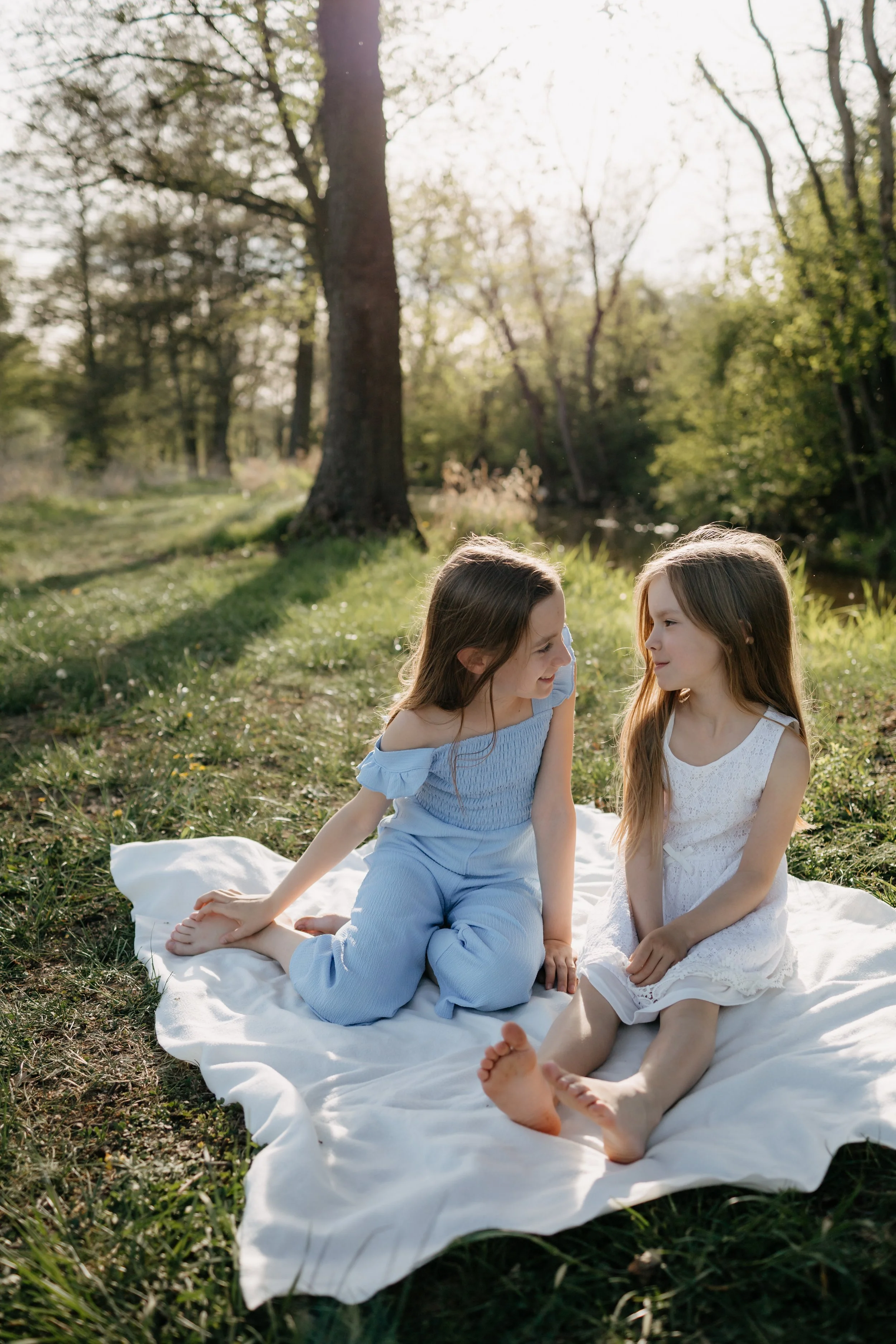 Two young girls sitting on a white blanket in a grassy area near a stream, surrounded by trees on a sunny day.