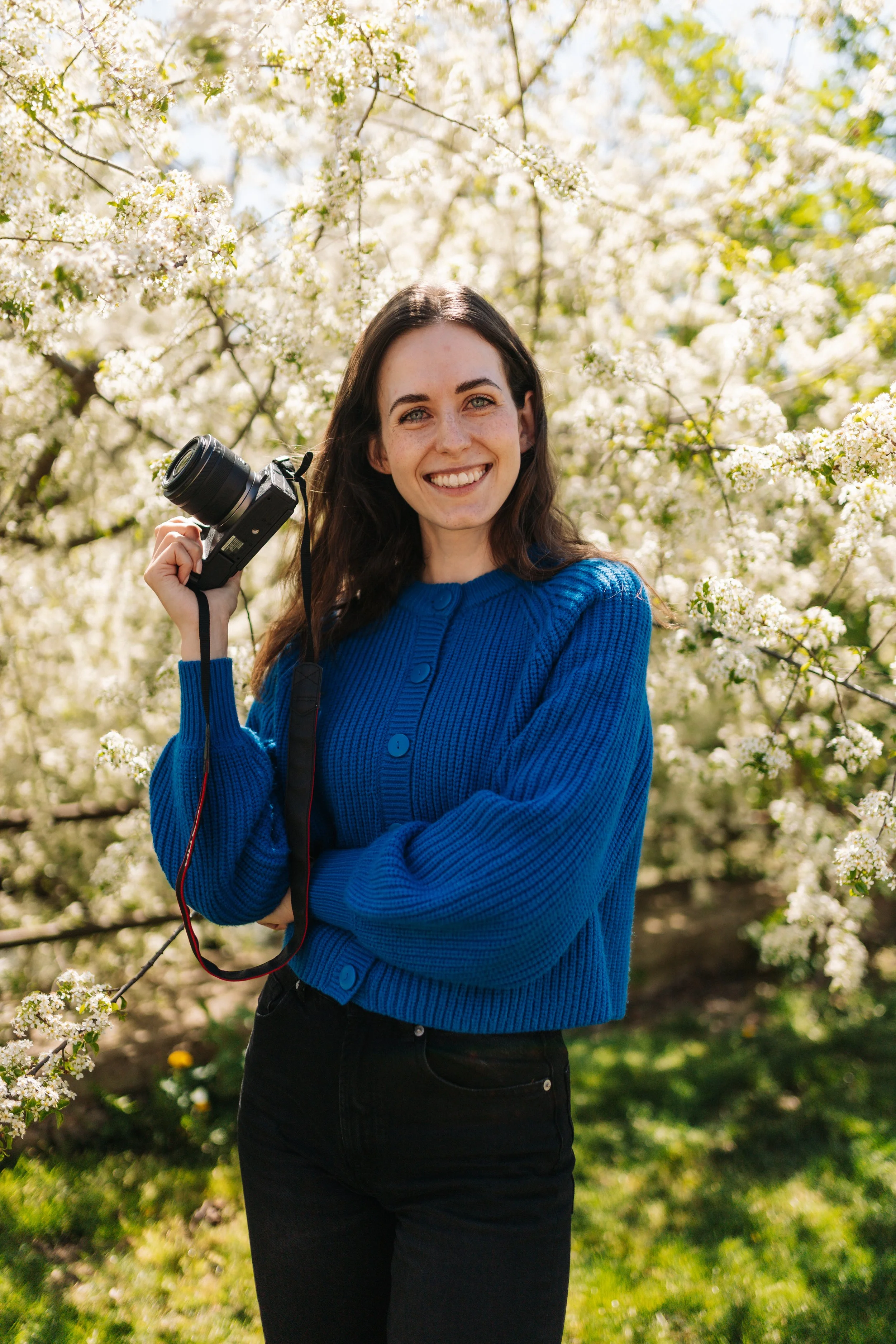 A young woman with long brown hair and a bright smile holding a camera, standing in front of blooming white flowers on trees on a sunny day.