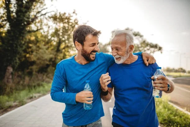 Two men, one young and one older, wearing blue shirts and holding water bottles, walking together outdoors and laughing.