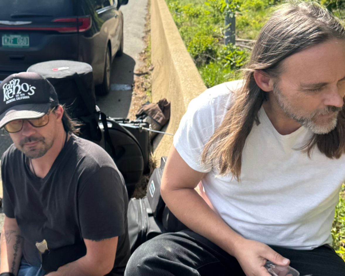 Two men sitting on the curb beside a parked car on a sunny day, with green trees behind them.