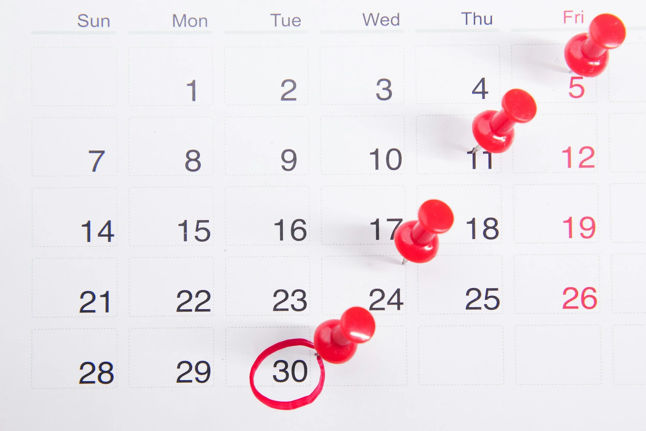 A calendar page showing the days of the week from Sunday to Saturday. Several red push pins are placed on dates within the calendar, specifically on the 5th, 11th, and 30th of the month.
