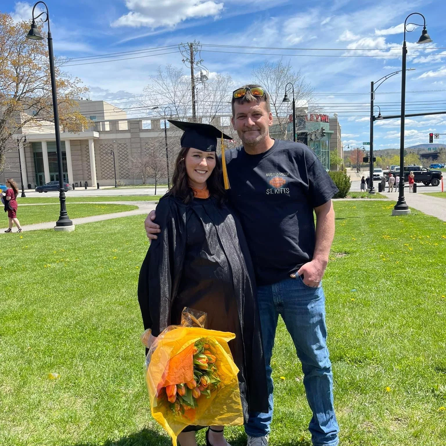A woman in a black graduation gown and cap, holding a bouquet of orange tulips, stands beside a man wearing a black Harley Davidson T-shirt, outdoors on a sunny day. They are smiling with their arms around each other. In the background, there are trees, street lamps, a building, and a few people walking.
