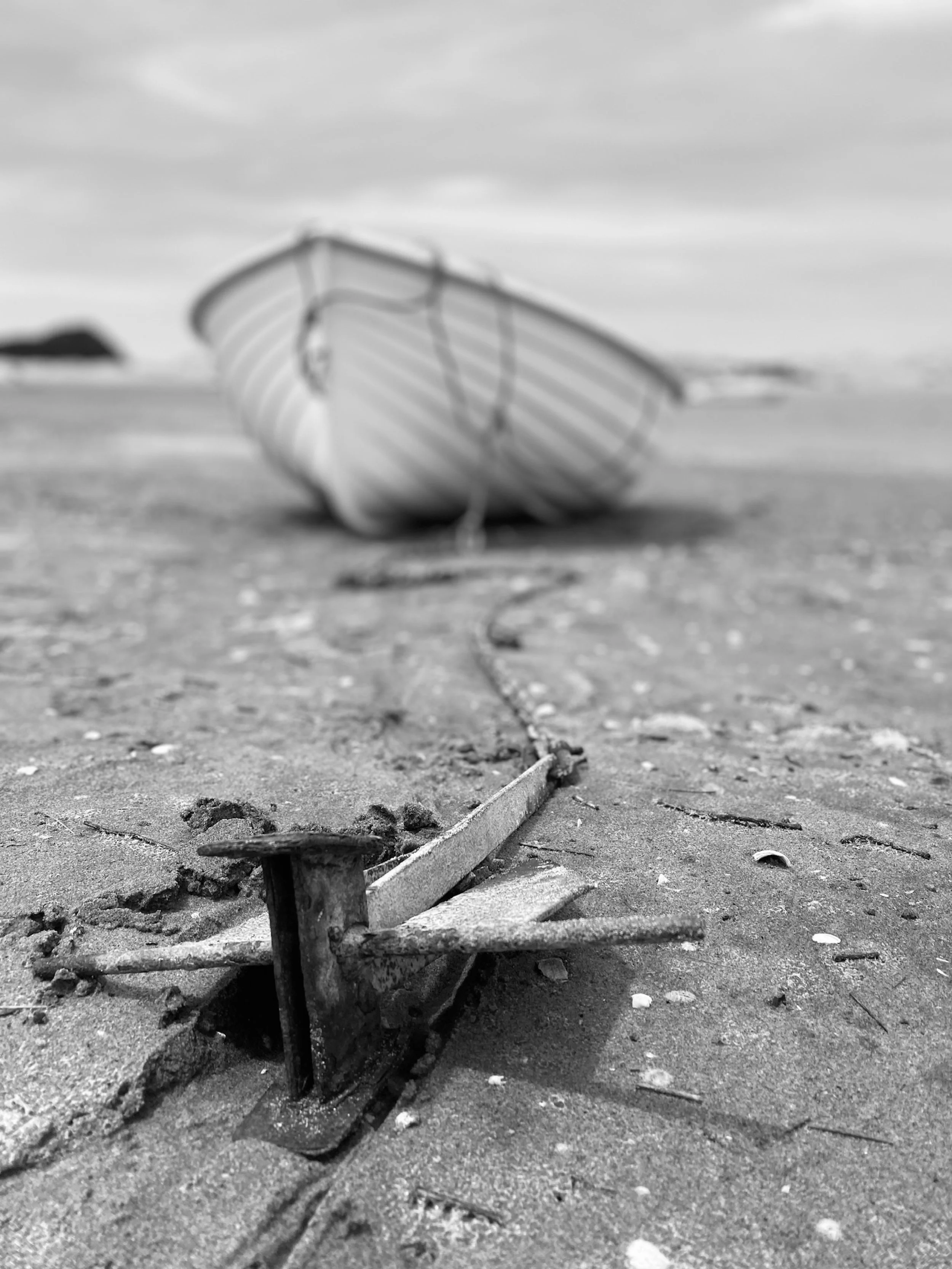 Close-up of a rusty, weathered nail and broken wood on a sandy beach with blurred boat in the background.