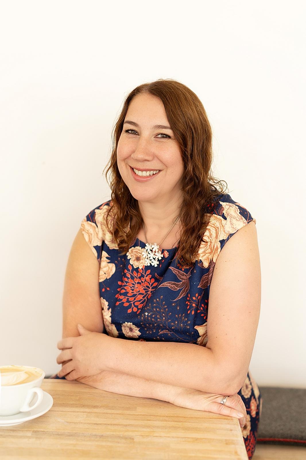 A woman with auburn hair smiling at the camera, seated at a wooden table with a cup of coffee.