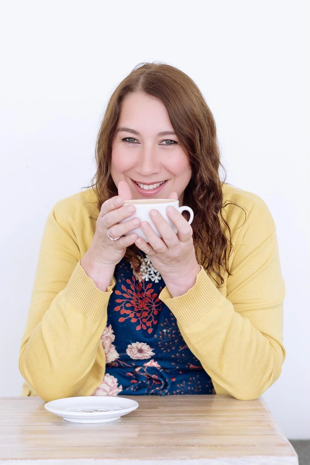 A woman with red hair smiling happily while holding a white cup, sitting at a table with a saucer in front of her, against a plain white background.