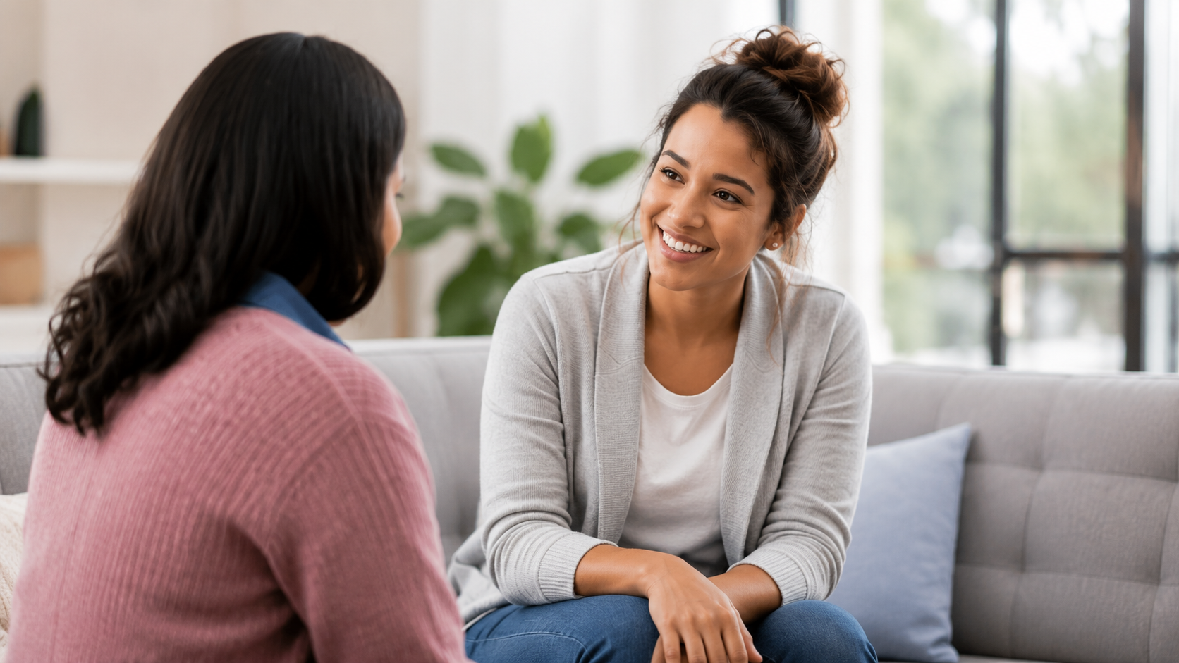 Two women having a conversation on a sofa in a bright, cozy room with a large window and houseplants.