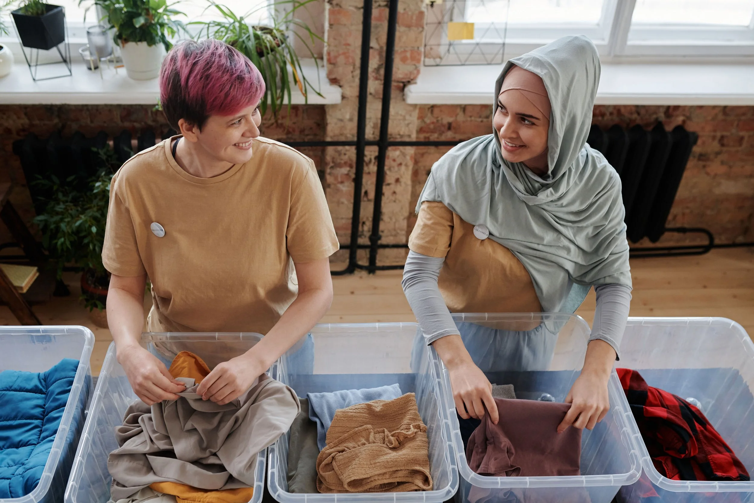 Two women with short hair, one with pink hair and the other wearing a hijab, are smiling and packing clothing into clear plastic storage bins in a room with brick walls and a large window.