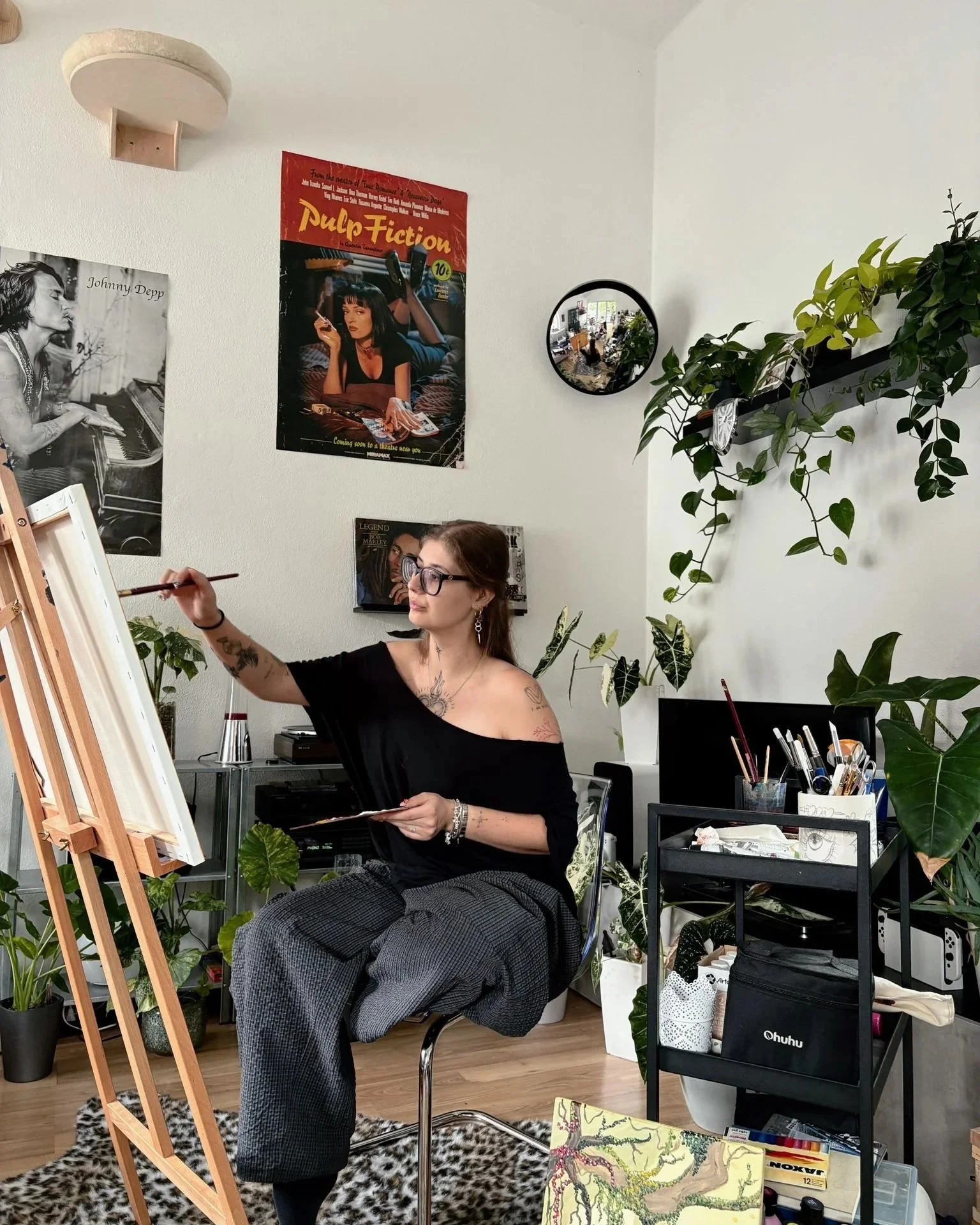 Eine Frau mit Tattoos sitzt im kunstvollen Raum, malt auf einer Staffelei, umgeben von Pflanzen, Postern und Künstlerbedarf, mit einem Spiegel, der den Raum reflektiert.