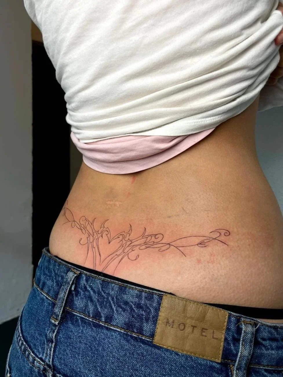 Nahaufnahme eines Bauches mit Tattoo eines stilisierten Baumes mit kleinen Zweigen, während die Person eine weiße und pinke Hemd trägt und eine Jeans mit Lederlabel."MOTEL".