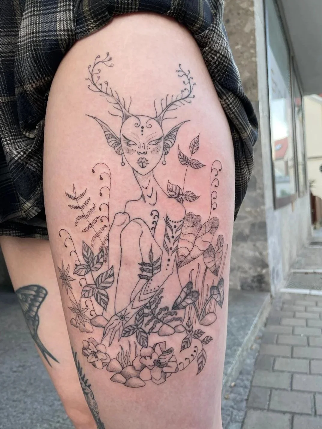 Lineart-Tattoo eines Elfen mit Geweih, umgeben von Pflanzen und Blumen auf einem Oberschenkel.