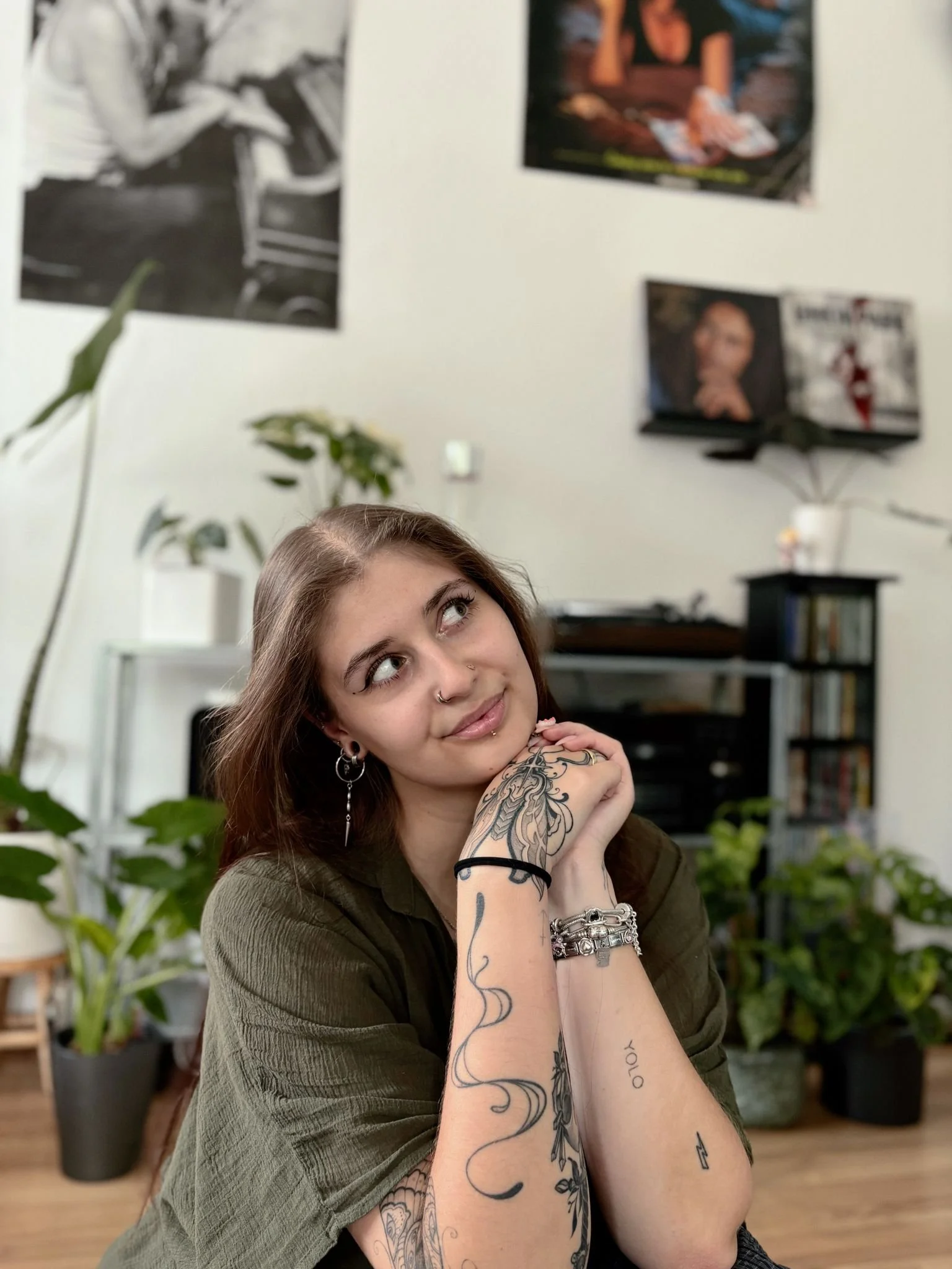Junge mit braunen Haaren, Arme voller Tattoos, sitzt in einem Raum mit Pflanzen und Plakaten an der Wand.