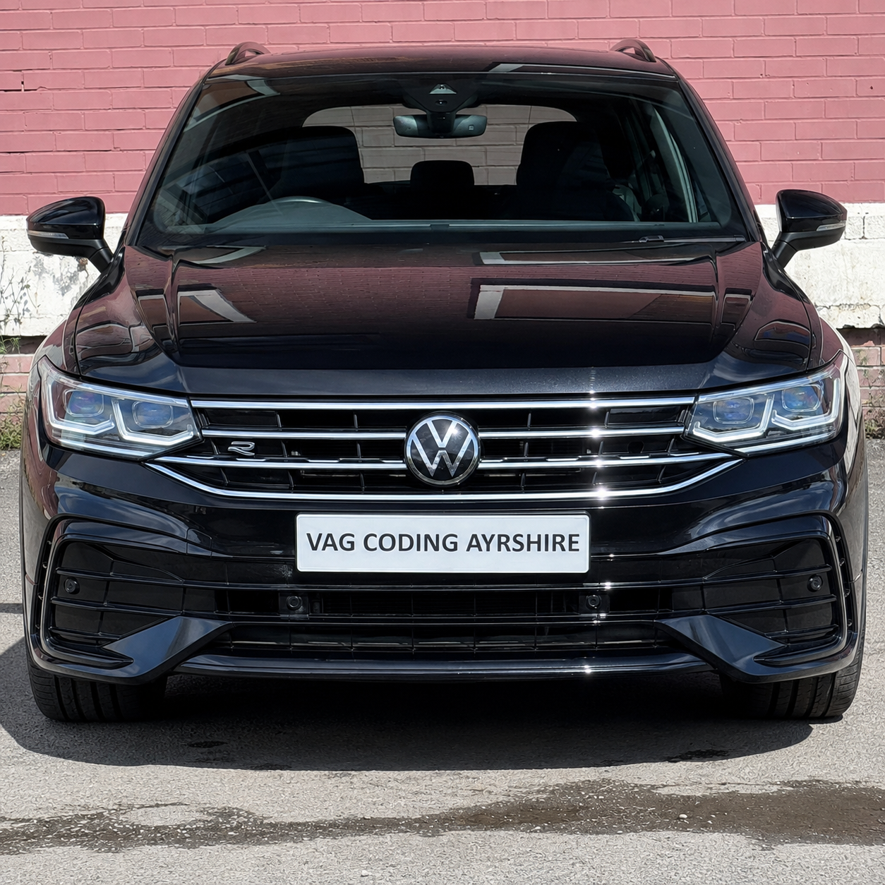 Front view of a black Tiguan MK2 Volkswagen car parked against a pink brick wall, with a white license plate that reads 'VAG CODING AYRSHIRE.'