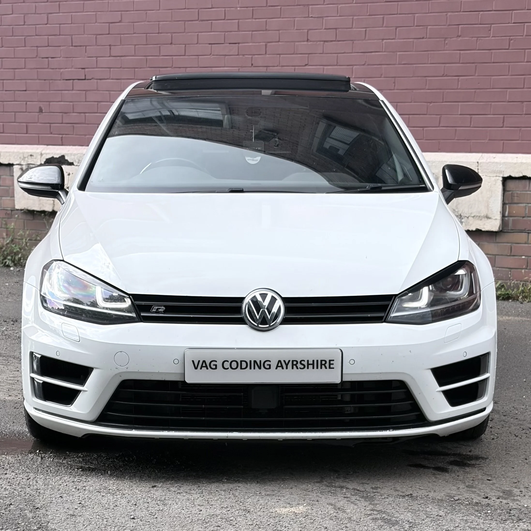 Front view of a white Volkswagen car parked on the street with a brick wall in the background.