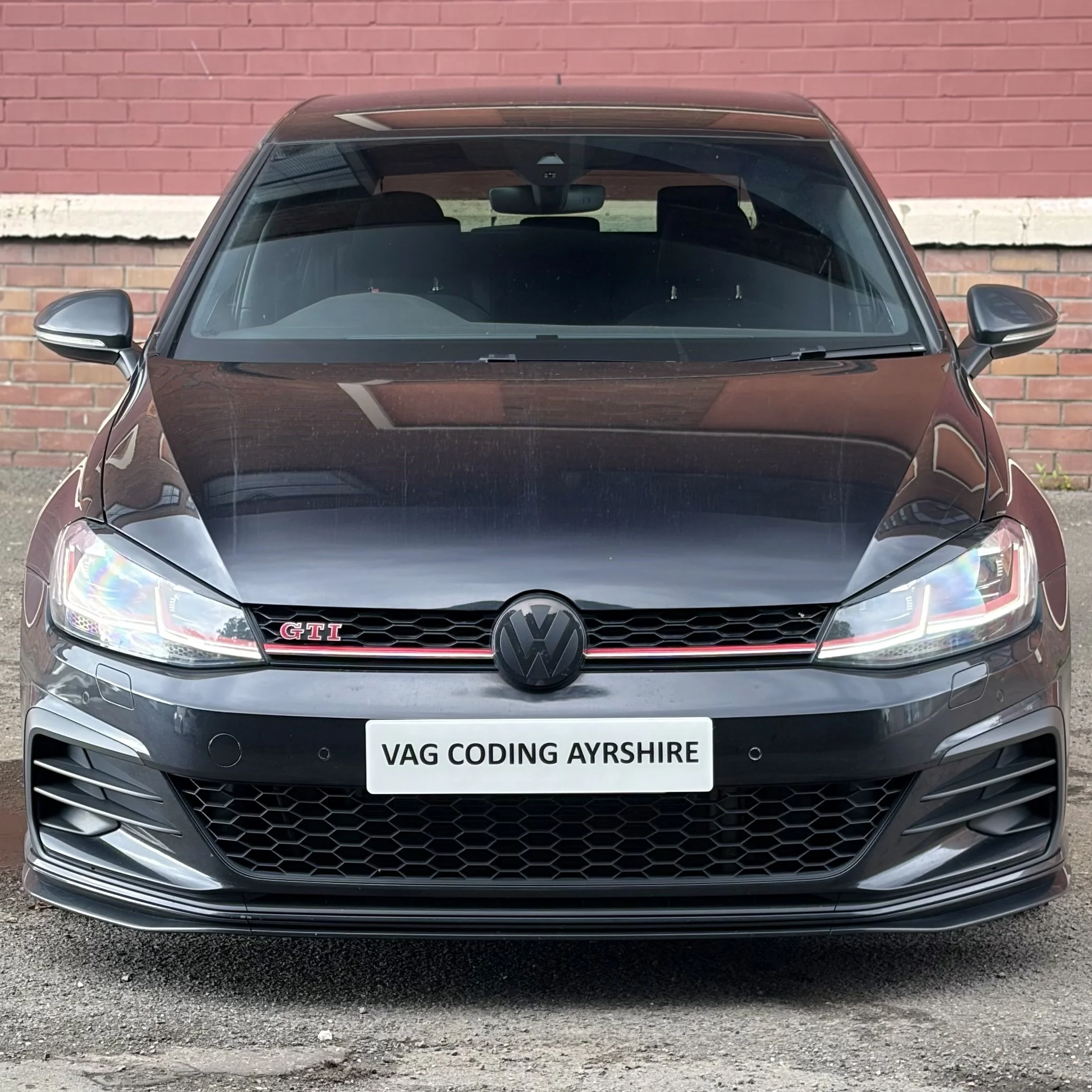 Close-up front view of a black Volkswagen GTI car parked in front of a red brick wall, showing the front grille, headlights, and side mirrors.