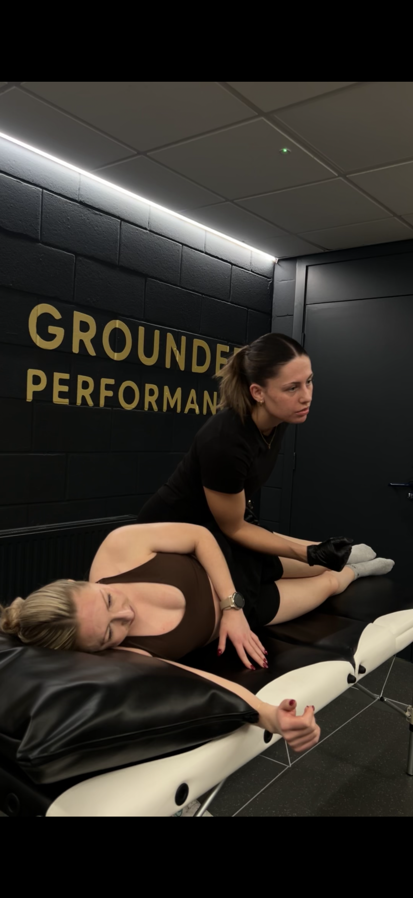 A woman lying face down on a massage table while another woman, wearing black gloves, performs a massage or medical treatment on her back in a room with black walls and the words 'GROUNDER PERFORMANCE' written on the wall.