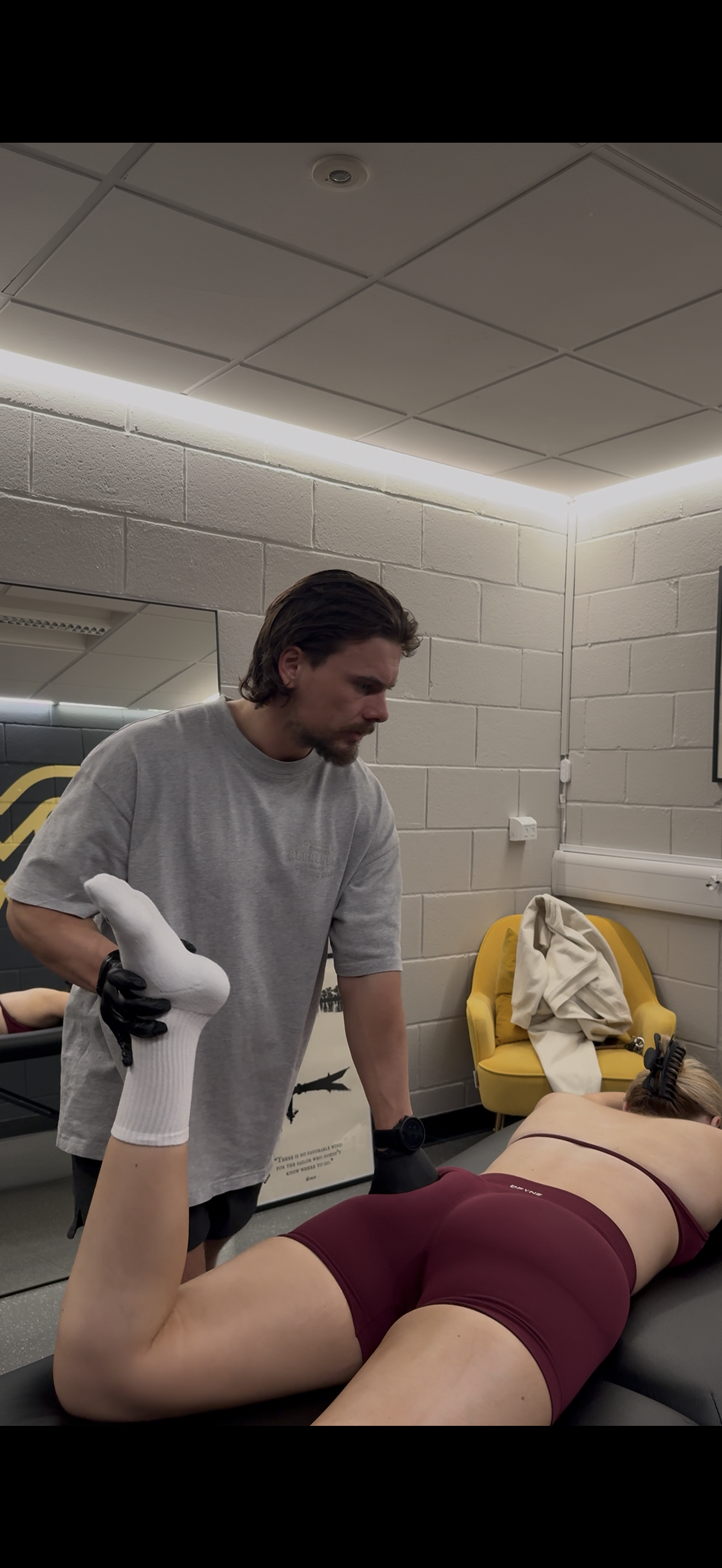A man with dark hair in a gray shirt is giving a massage or therapy to a woman lying face down on a massage table. The woman is wearing maroon workout shorts and a sports bra, with her arm raised and hand in a white glove holding a device against her lower back. The room has beige brick walls, a yellow chair with a beige cloth on it, and a mirror on the wall.