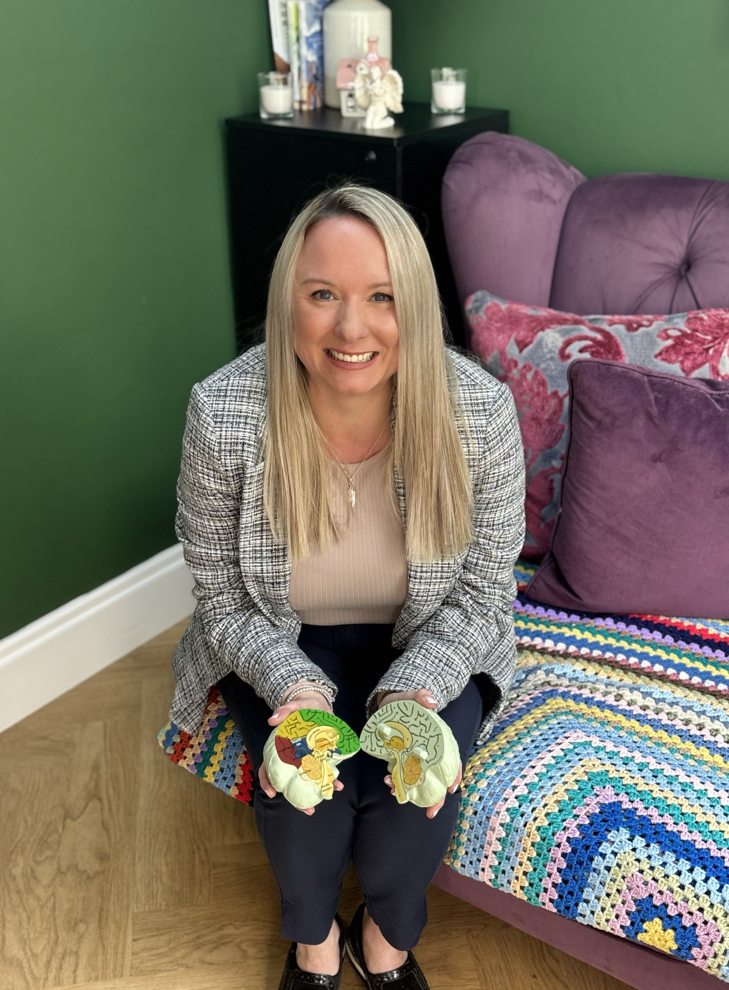 A woman with long blonde hair, smiling, seated on the floor in a living room, holding brain model halves, wearing a plaid blazer and black pants, with purple cushions and multicolored blanket on a purple velvet sofa behind her, and a black cabinet with candles and decorative items against a green wall.