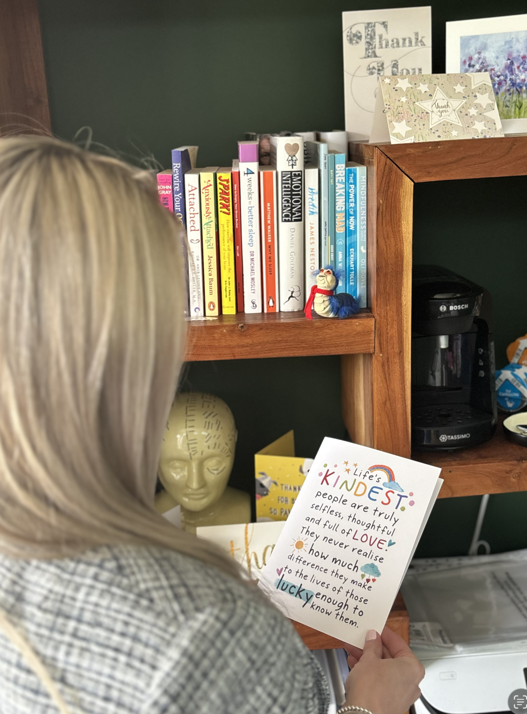 Person holding a colourful card with a motivational message, standing in front of a bookshelf filled with books and decorations, including a small stuffed animal.