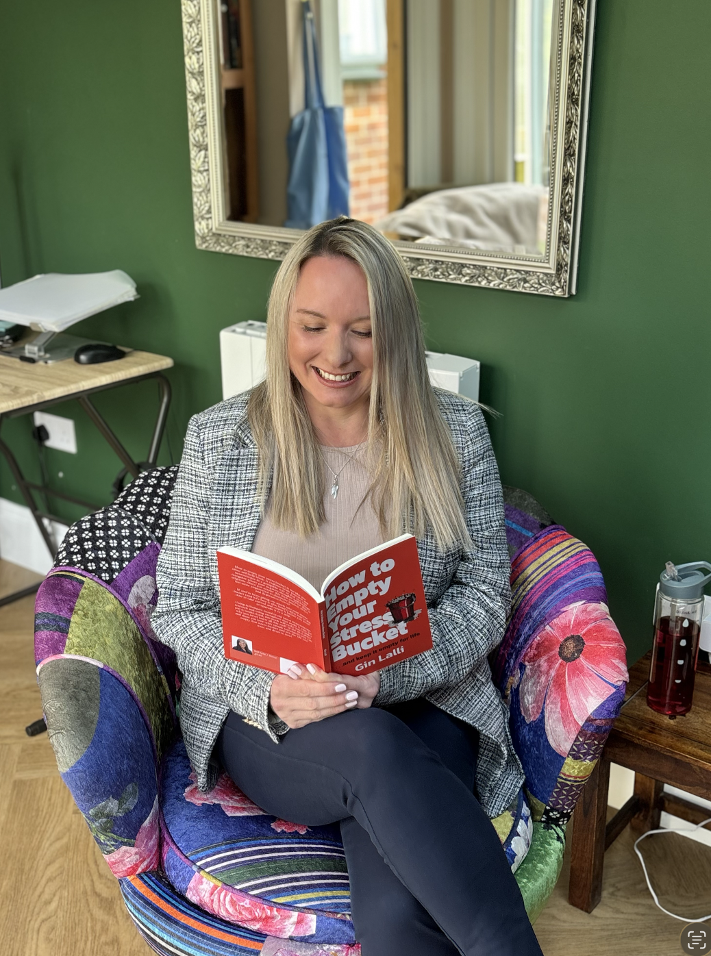 A woman with blonde hair smiling and reading a red book titled "How to Empty Your Stress Bucket" in a colourful floral armchair in a room with green walls, a mirror, and a wooden side table.