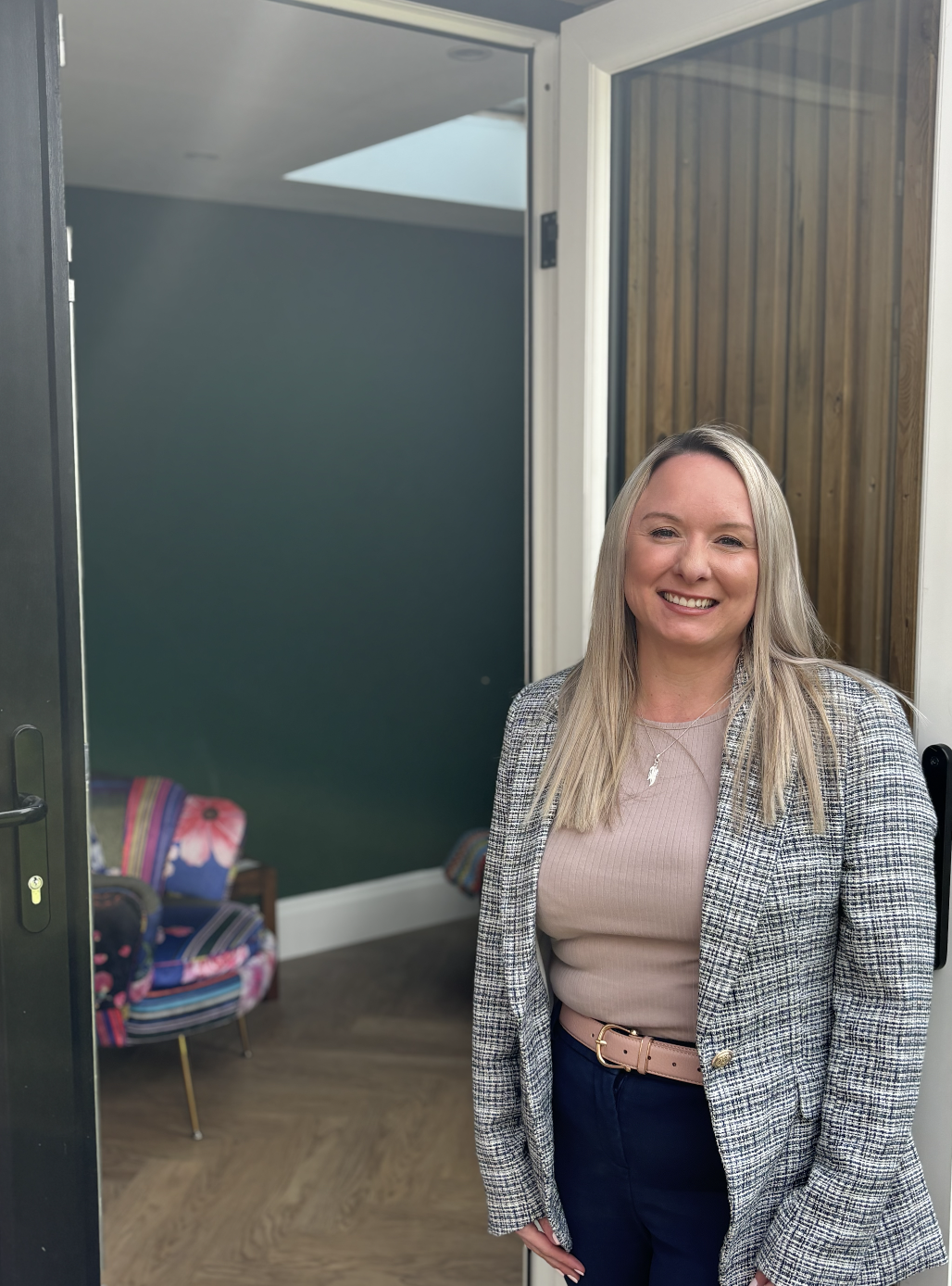 A smiling woman with blonde hair standing at an open door, with a colourful chair and a dark green wall in the background.