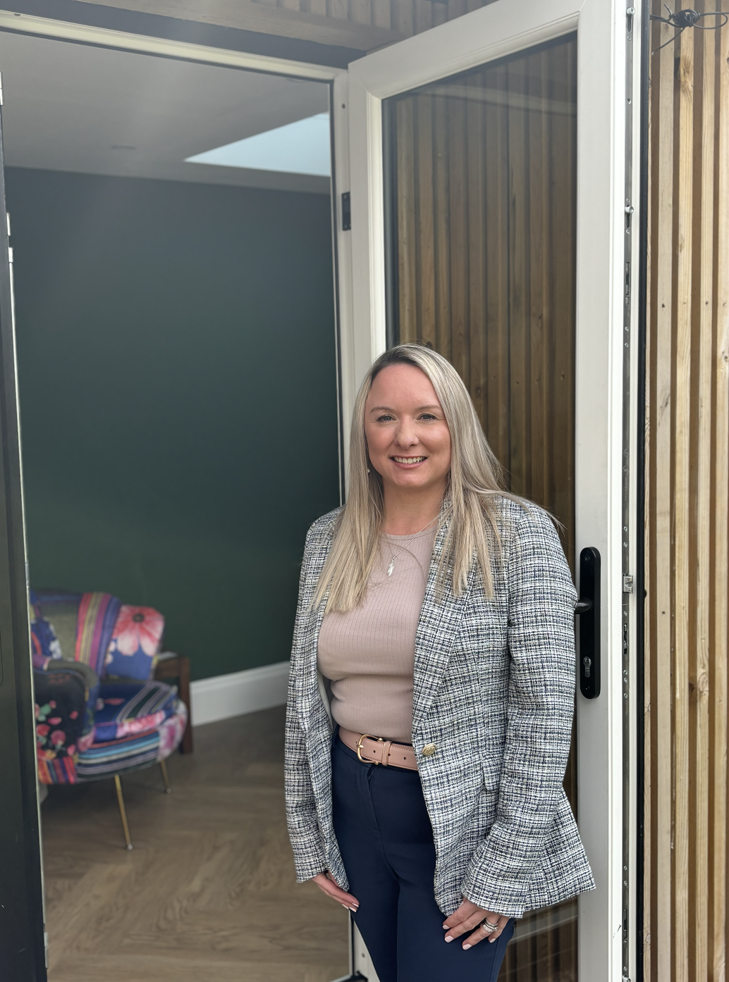A woman with blonde hair smiling and standing in front of a door opening into a room with green wall and colourful furniture.
