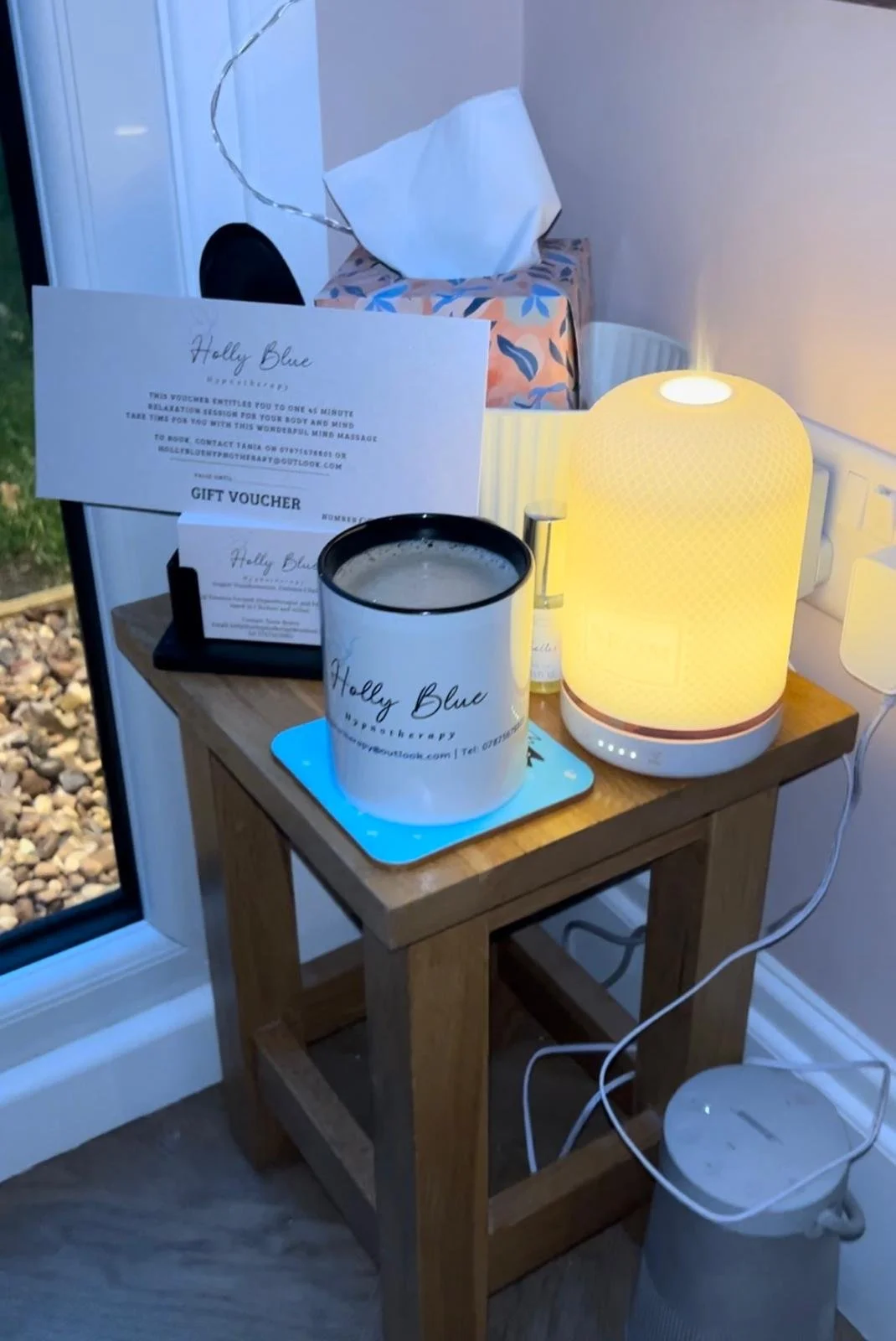 A wooden side table next to a window. The table holds a large white mug with black interior, a small box with gift vouchers and cards, a white diffuser with a yellow glowing lamp, a small spray bottle, and a cord plugged into a power outlet.