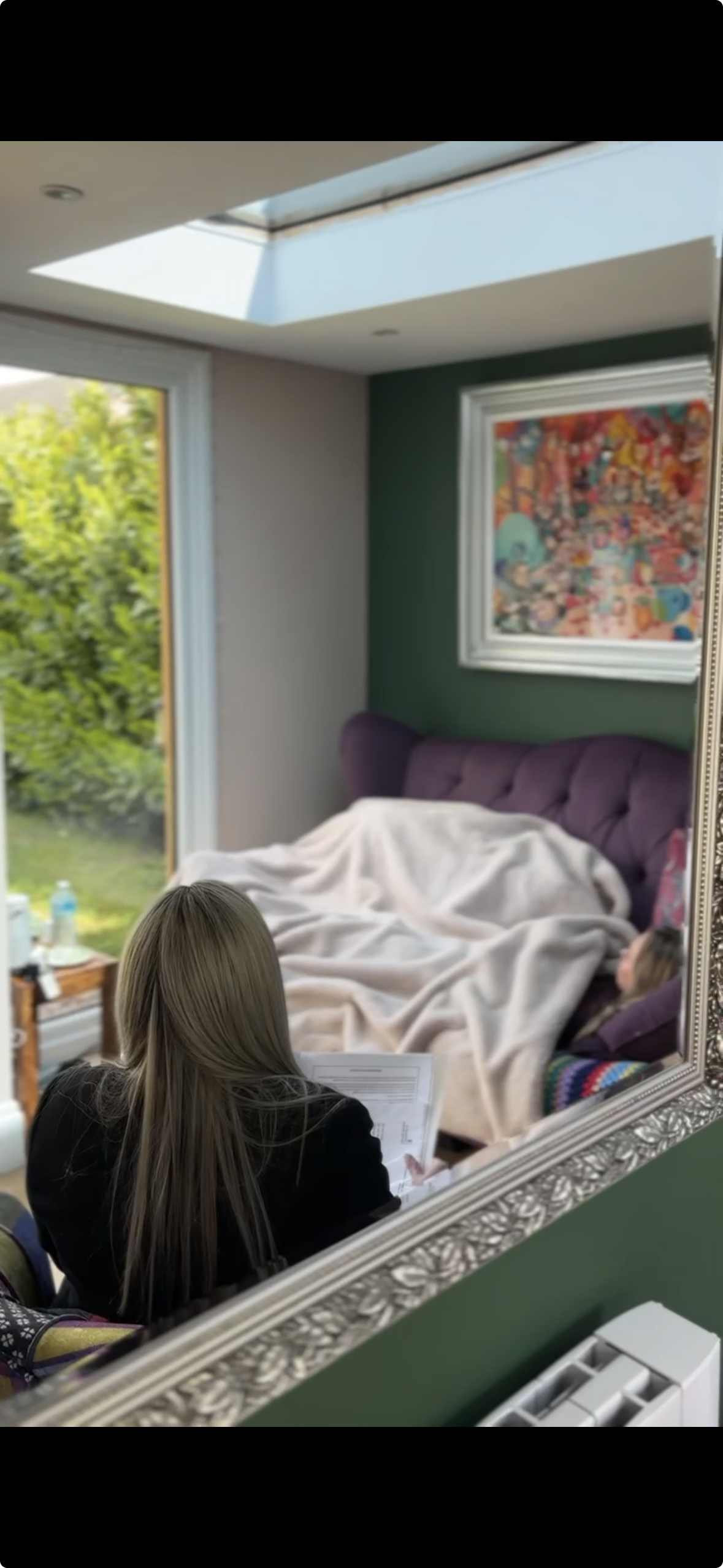 A person with long blonde hair sits in front of a large mirror, reviewing documents, while another person reclines on a purple sofa with a beige blanket, in a room with a green accent wall, colourful artwork, and a skylight.