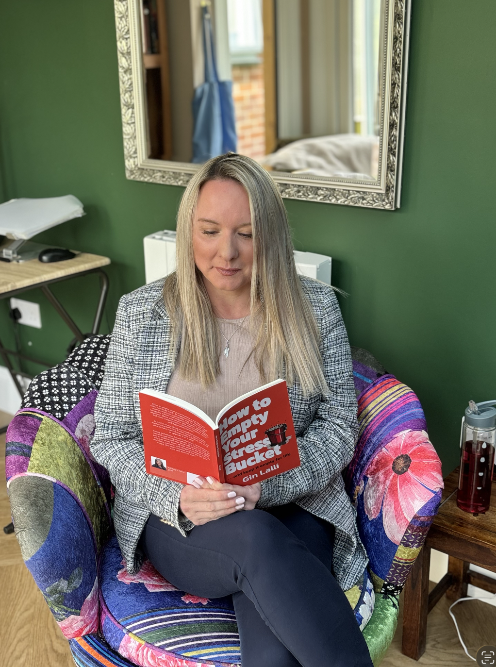 A woman sitting in a colourful armchair reading a book titled 'How to Empty Your Stress Bucket' in a room with green walls, a large decorative mirror, and a window with curtains.