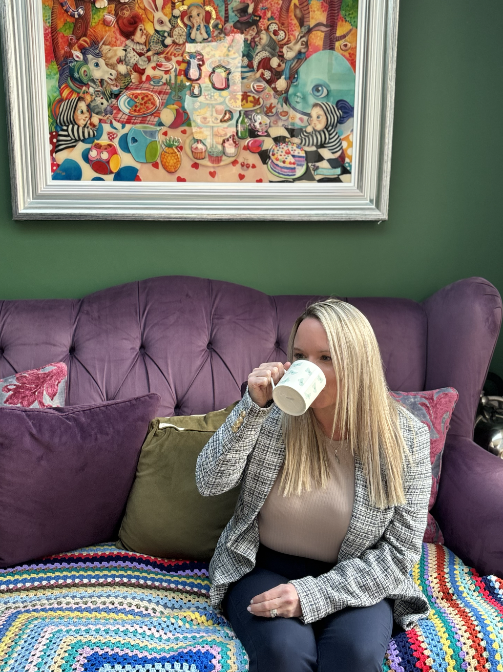 Woman with blonde hair sitting on a purple velvet sofa, drinking from a white mug with a green pattern, in a room with a colourful painting on local green wall.